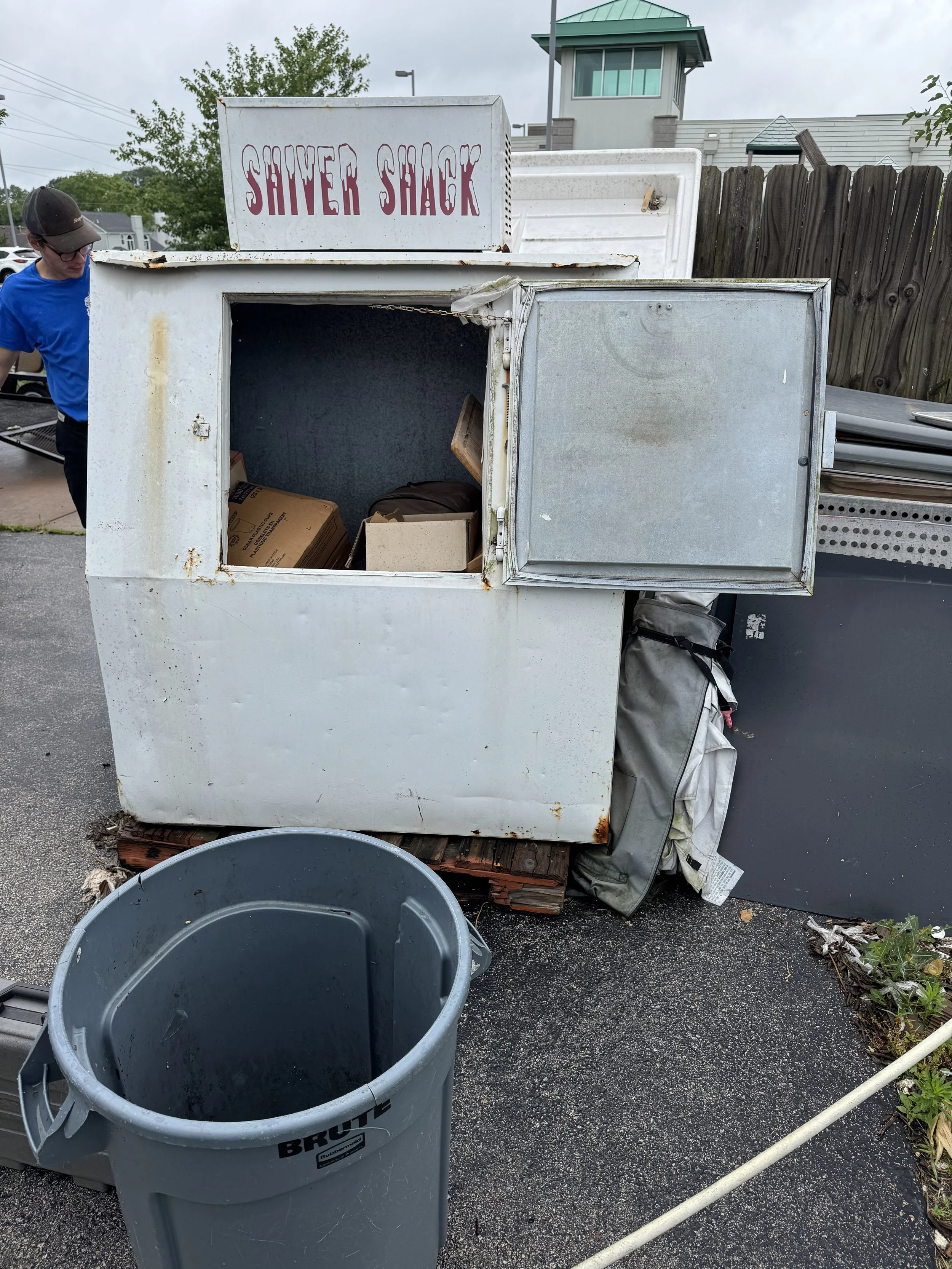 An outdoor scene featuring a weathered white ice shaver stand labeled 'Shaver Shack,' with a hinged metal door, several cardboard boxes inside, a gray trash bin labeled 'Brute' in front, and a person wearing a blue shirt and baseball cap partially visible to the left. In the background, there is a wooden fence, buildings, and a cloudy sky.