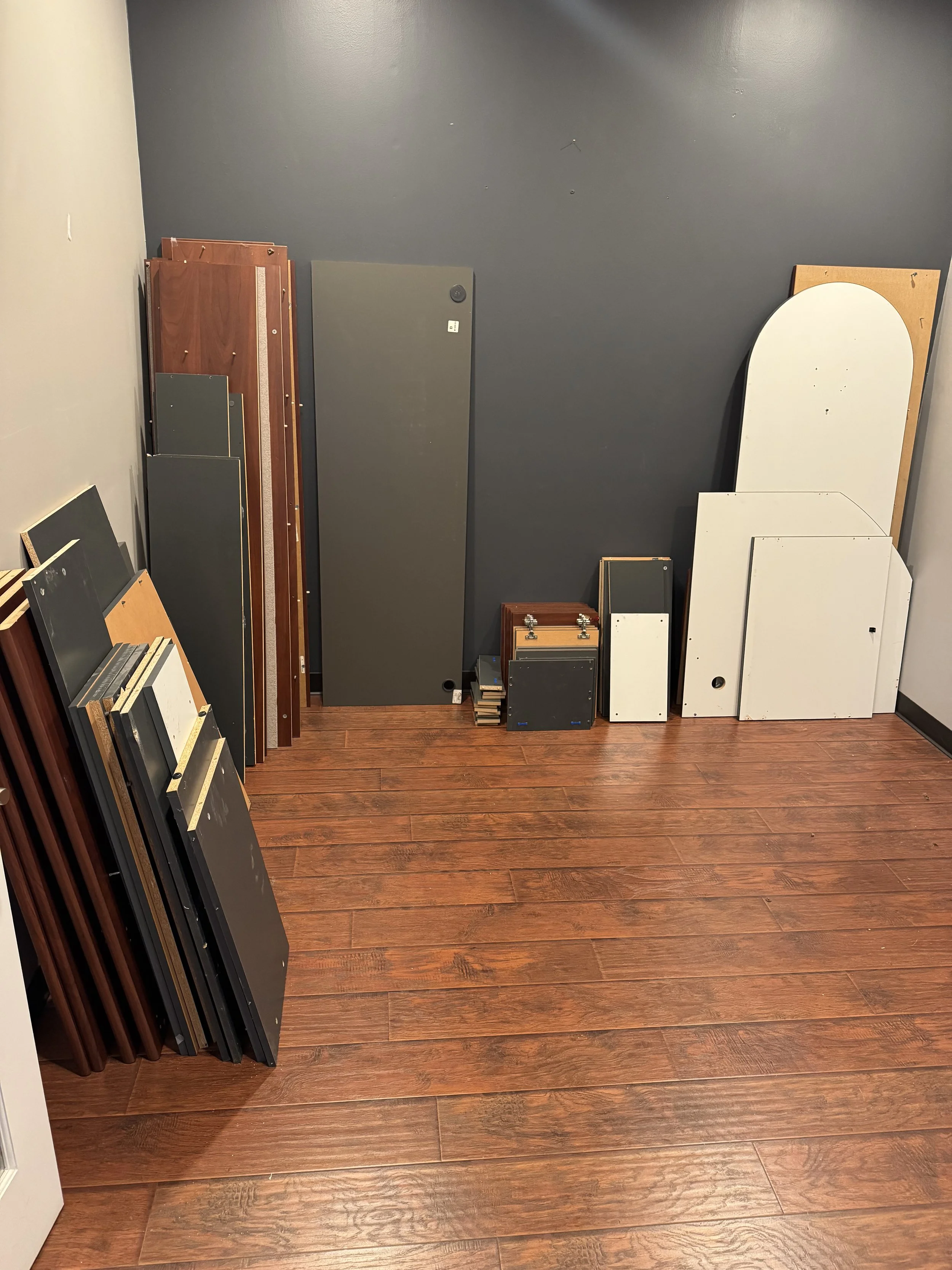 Room with wooden floor and gray and white walls, containing various disassembled furniture pieces, including cabinet doors, panels, and a small wooden frame with hinges, stacked and leaning against the wall.