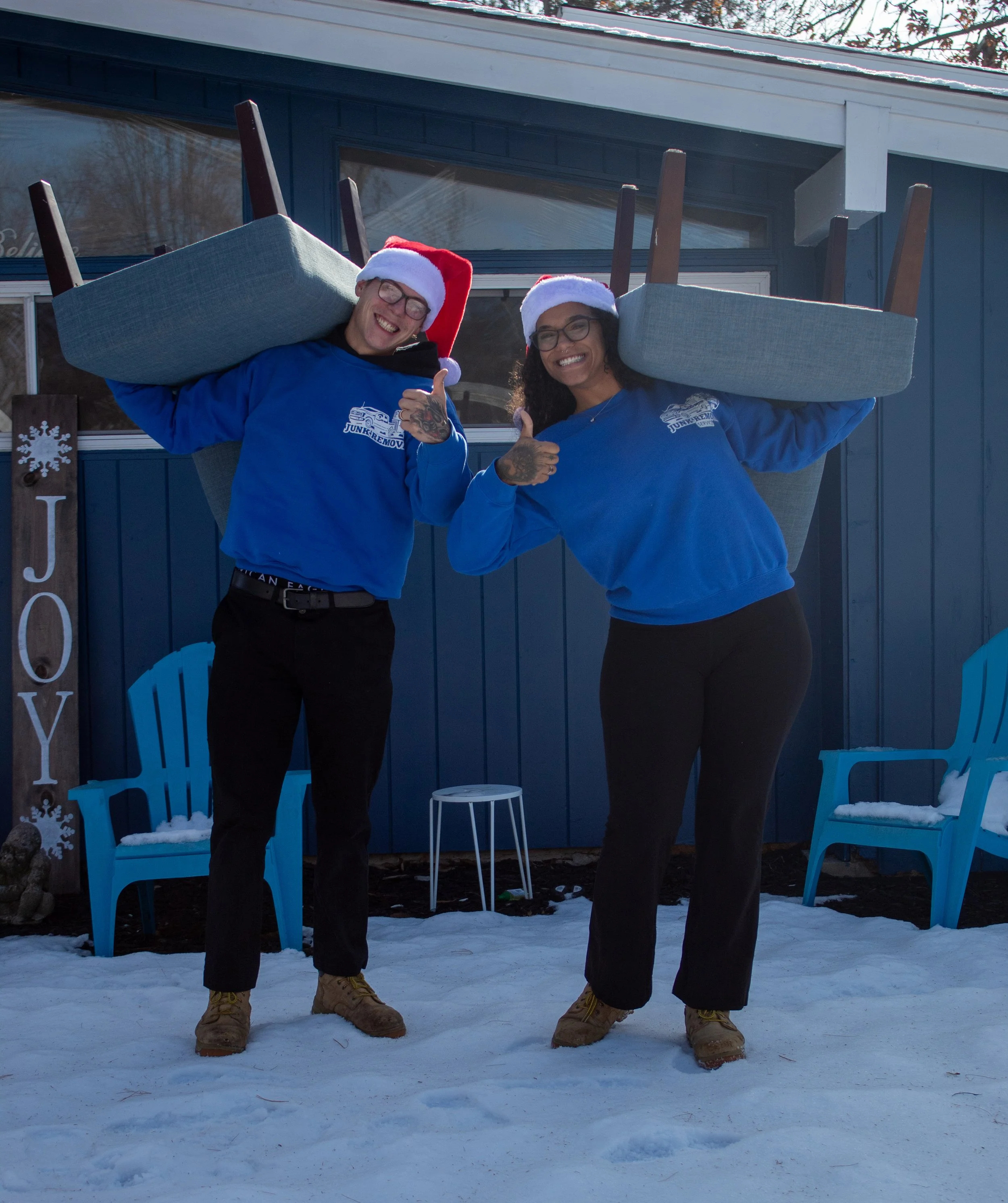 Two people wearing Santa hats and blue sweatshirts, carrying chairs on their shoulders, smiling and giving thumbs-up in front of a blue building with outdoor seating and snow on the ground.