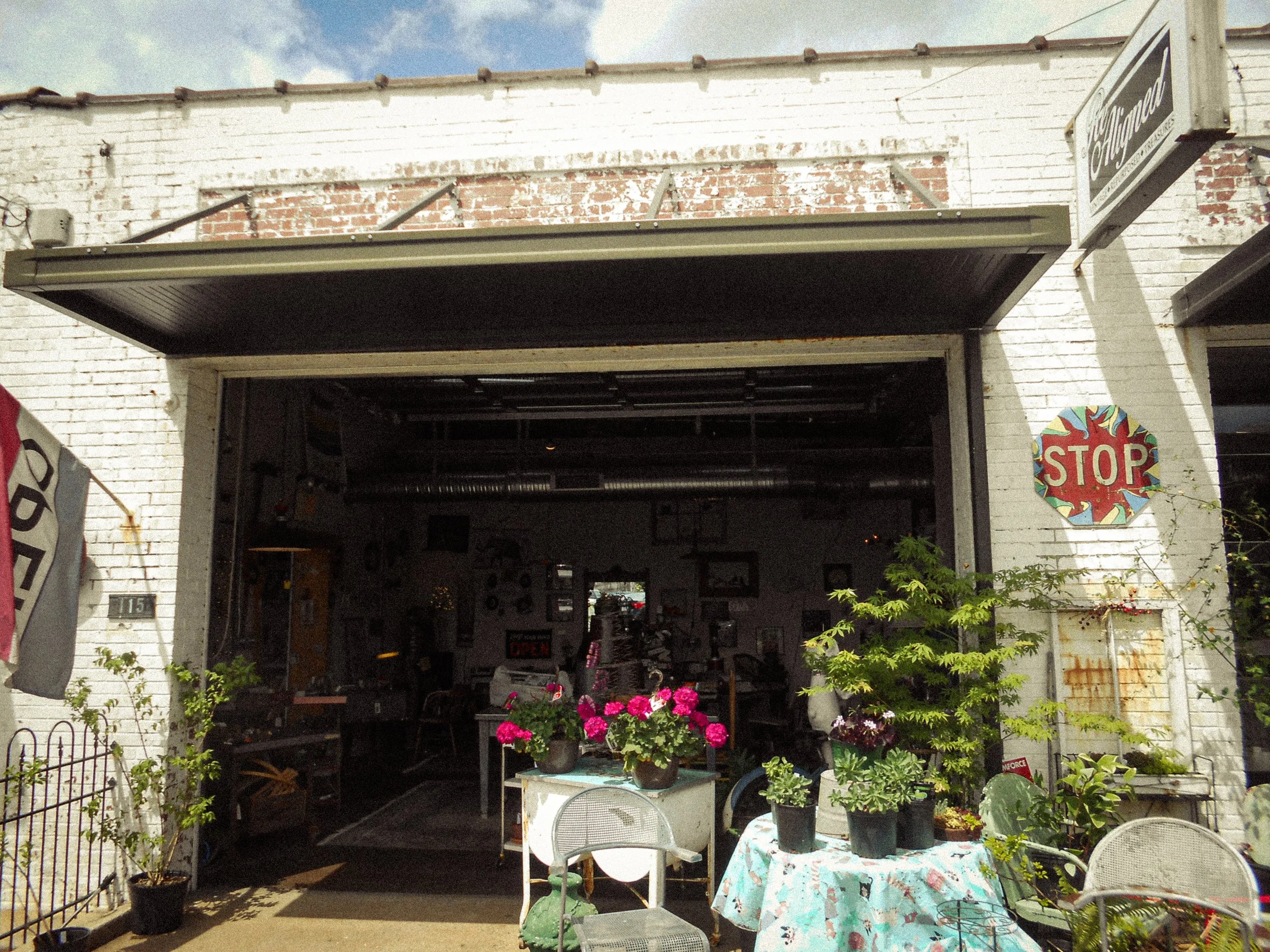 The front of a vintage shop with a partially opened garage door, displaying various items inside. Outside, there are potted plants, a small table with a floral tablecloth, and a couple of chairs. A colorful STOP sign is mounted on the wall near the door.