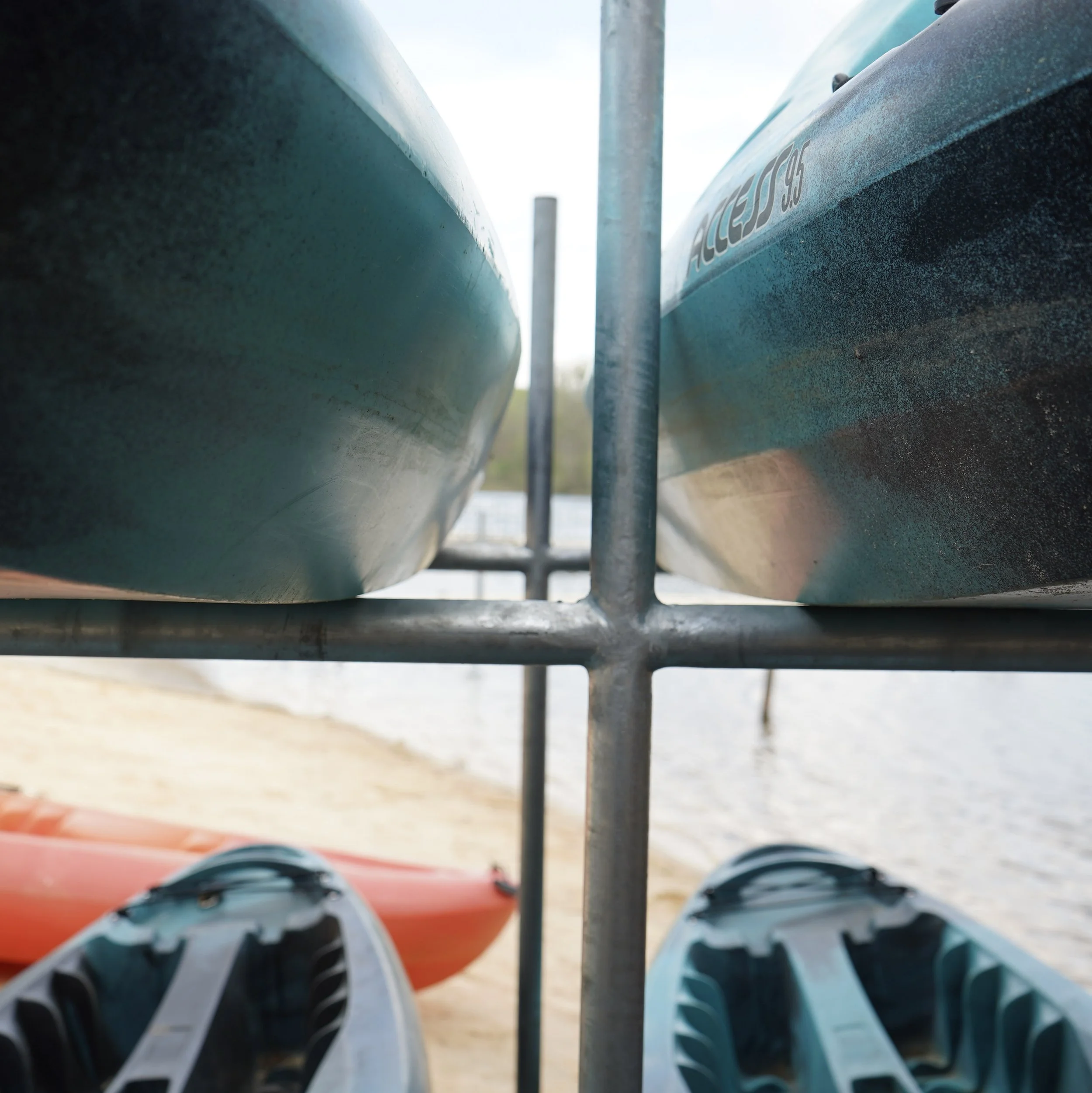 Close-up of two kayaks on a rack by the water, with a red kayak beneath two blue ones, viewed from a low angle.