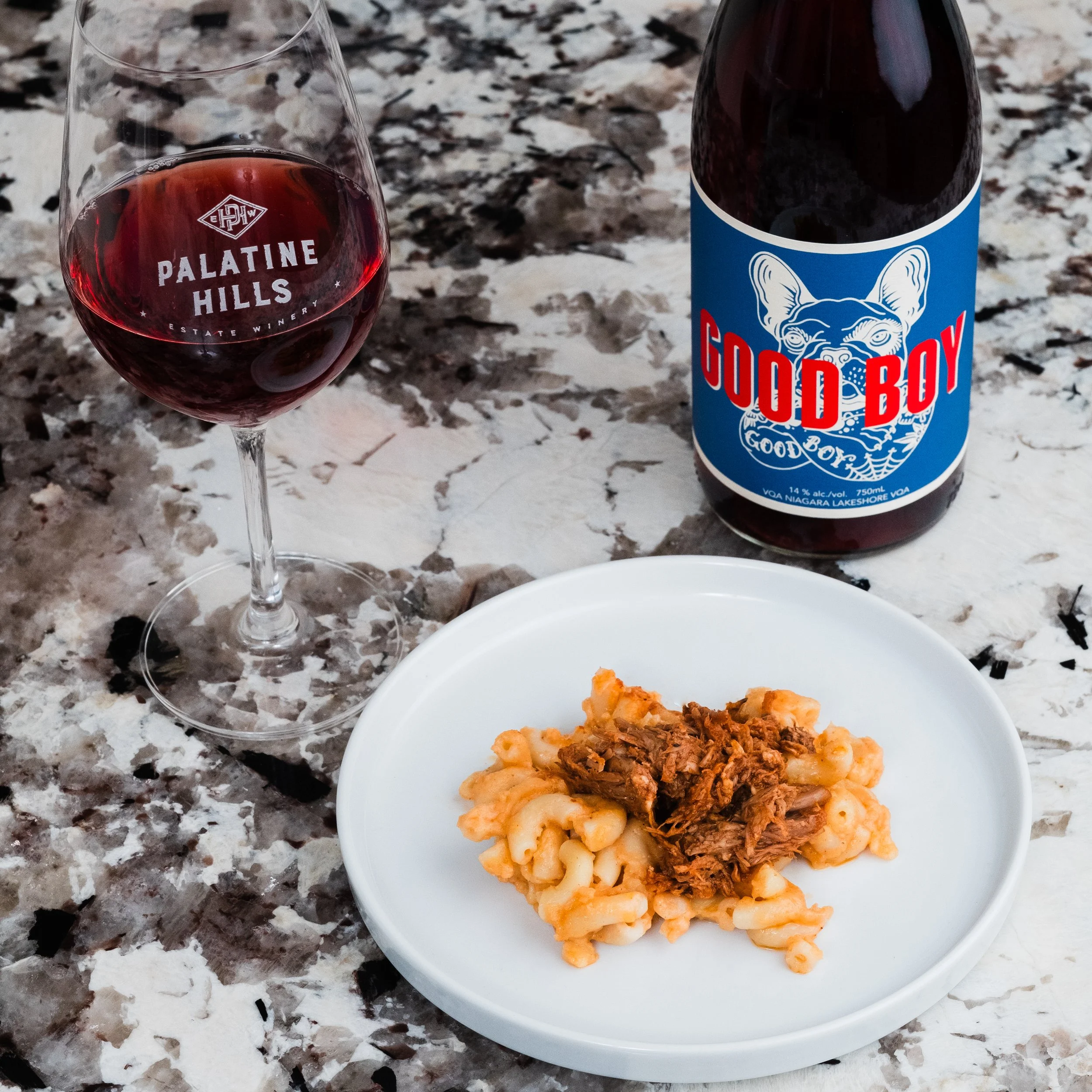 A plate of mac and cheese with pulled pork on top and a bottle of Palatine Hills Estate Winery Good Boy red wine