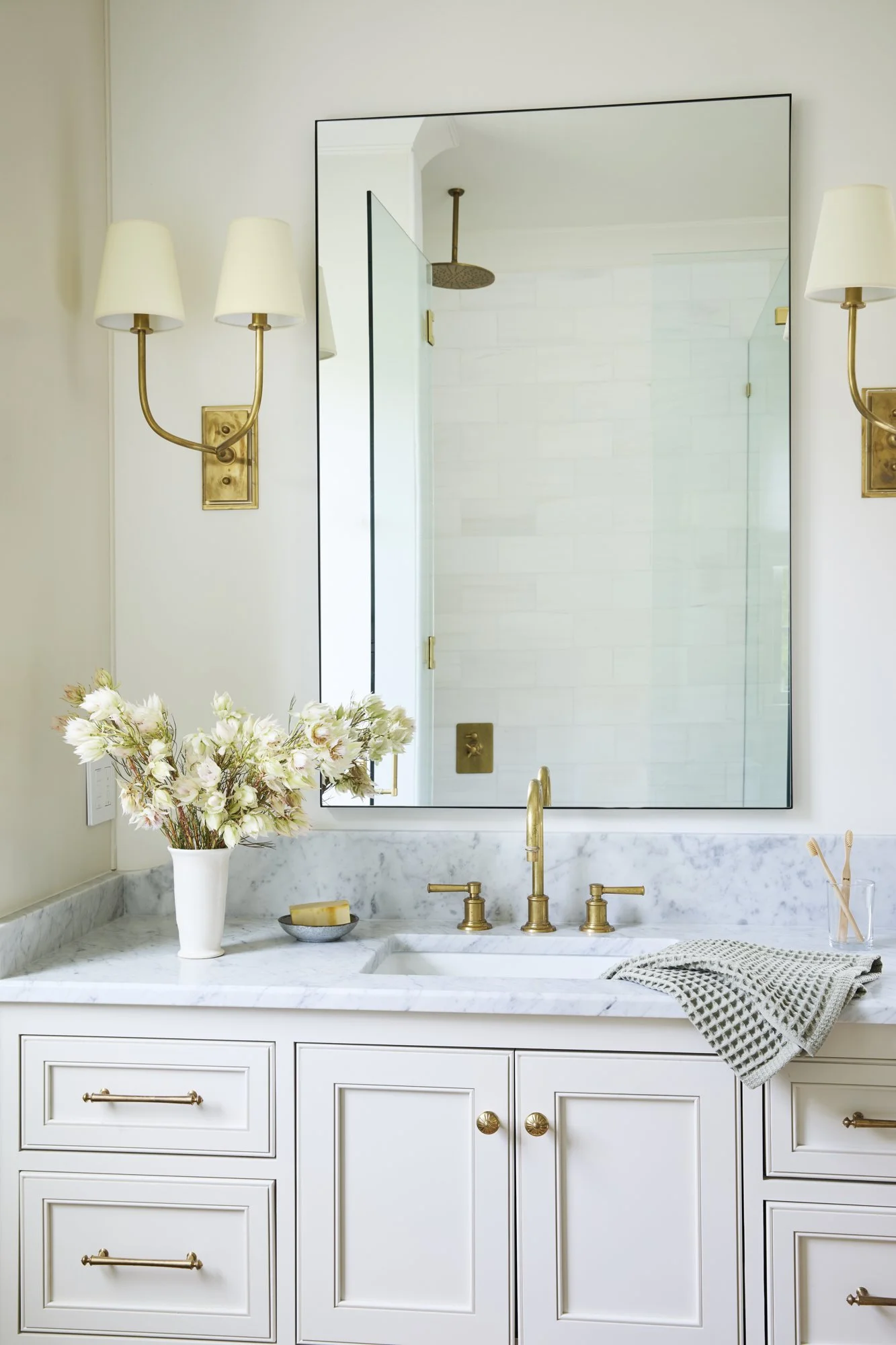 Bathroom vanity with a large mirror, white marble countertop, brass fixtures, a vase with flowers, soap dish, and toothbrush holder.