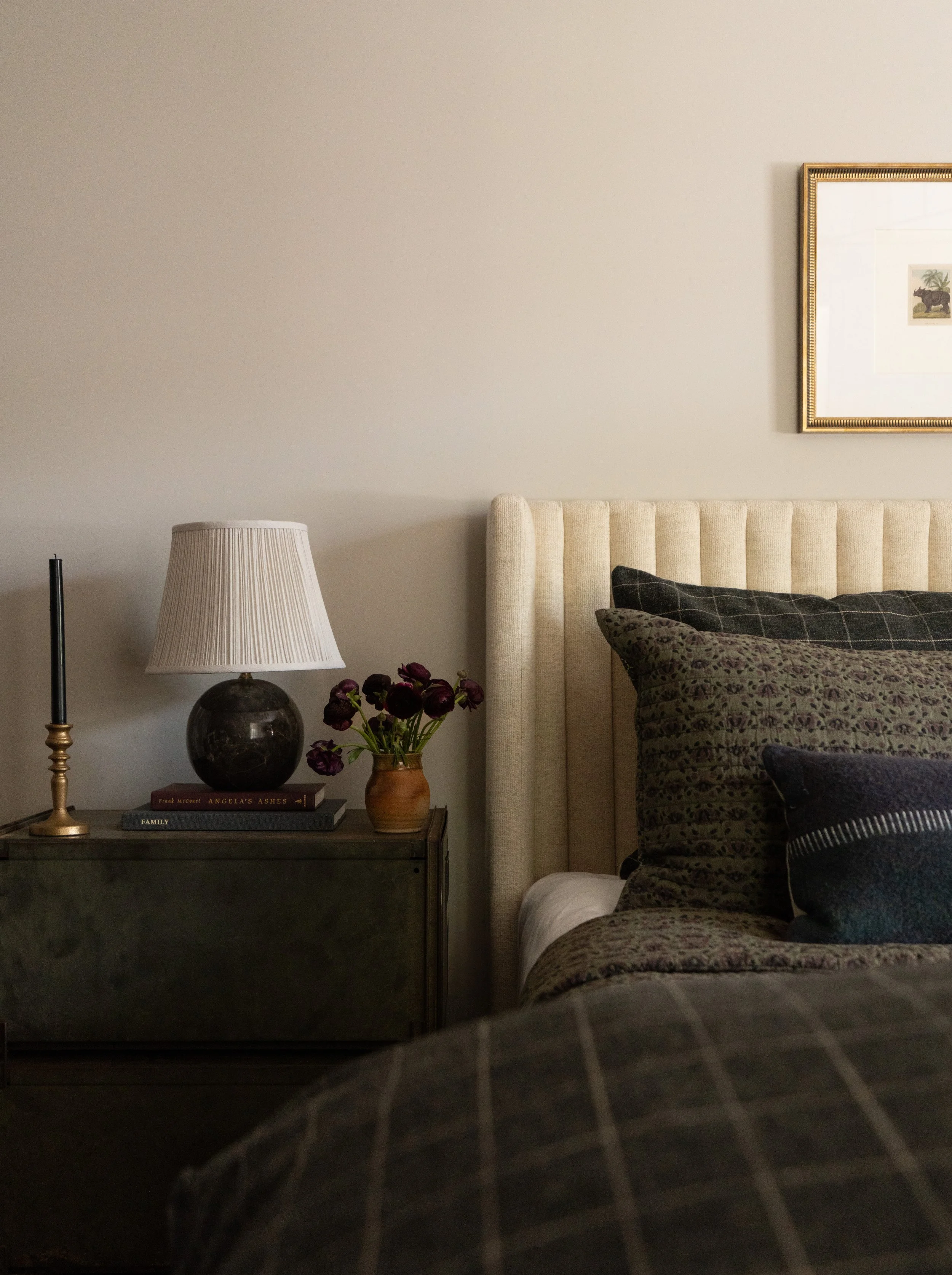A bedroom corner with a beige upholstered headboard, pillows, a dark wooden nightstand, a lamp with a pleated shade, a vase with dark purple flowers, and framed artwork on the wall.