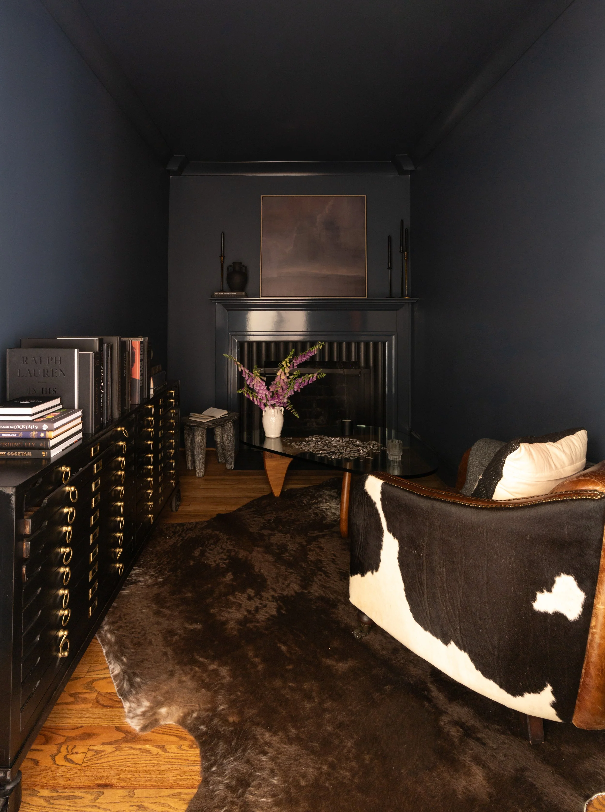 A cozy living room with dark navy walls, a black fireplace, a glass coffee table, a cowhide armchair, a black dresser with books, a vase with purple flowers, and framed artwork, all illuminated softly.