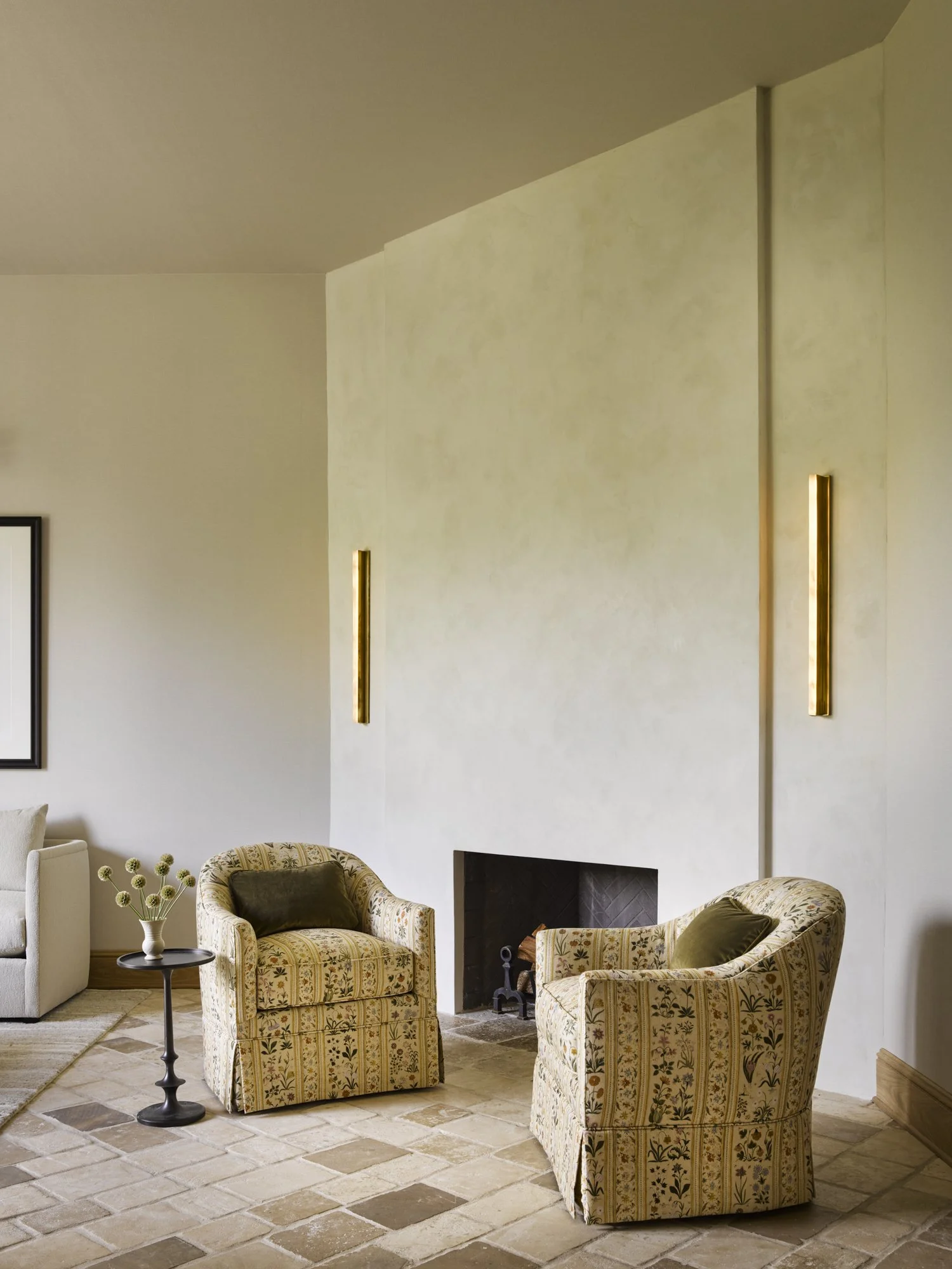 Living room with stone tile floor, two patterned armchairs, a small round table with a white vase and greenery, fireplace, and minimalist decor.