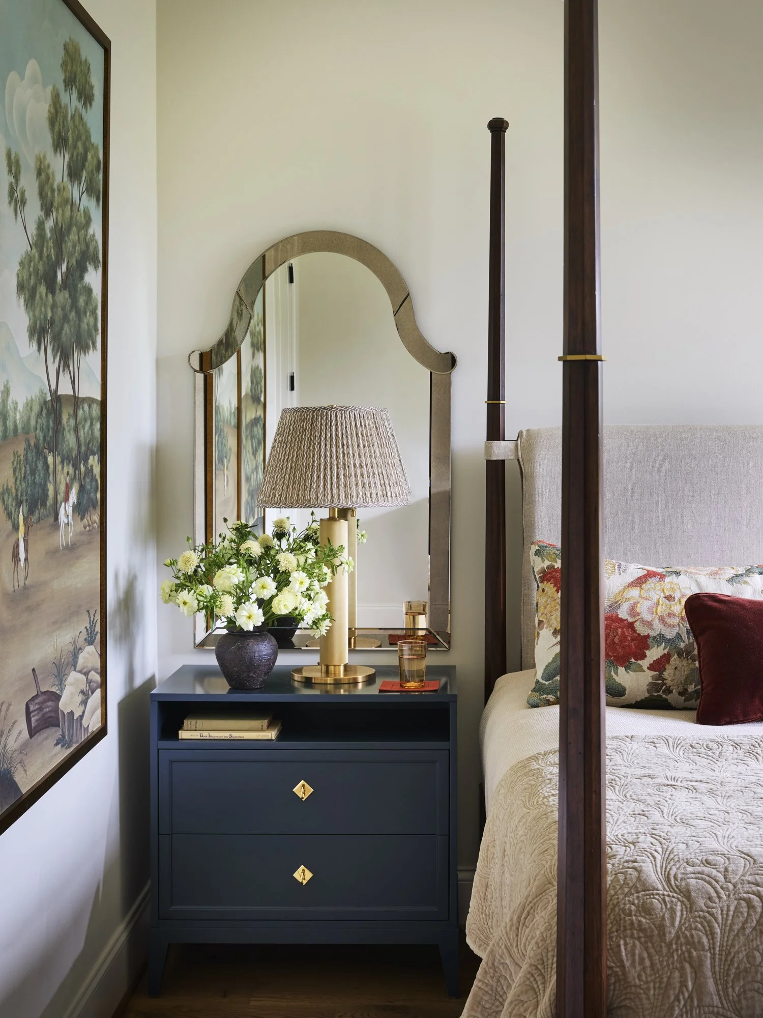 A bedroom corner with a beige bed and floral pillows, a navy blue nightstand with gold handles, a table lamp, a mirror, a vase with white flowers, a glass, and a decorative tray.