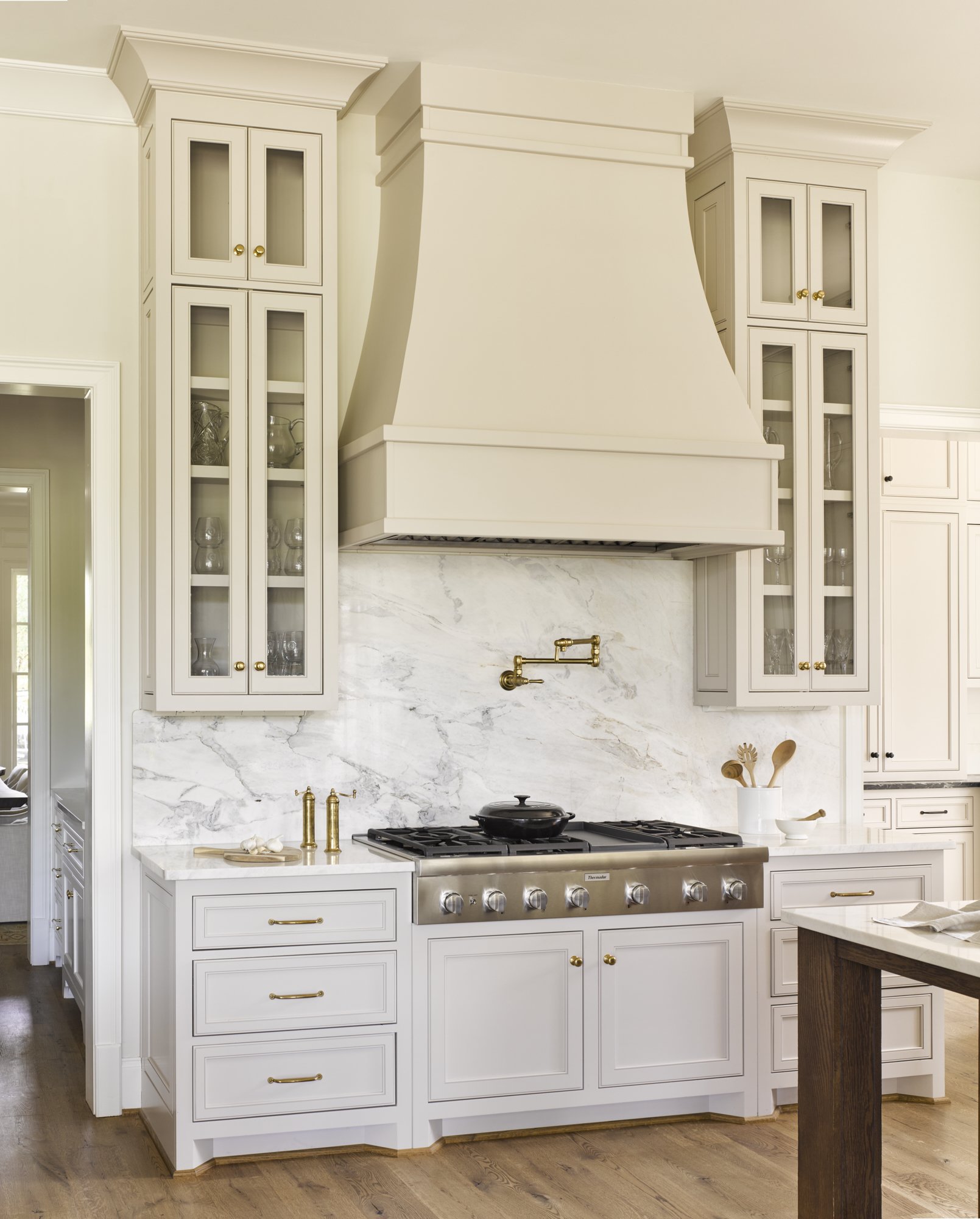 White kitchen with marble backsplash, brass fixtures, and a stainless steel stove. Cabinets above and below, with a wood floor.