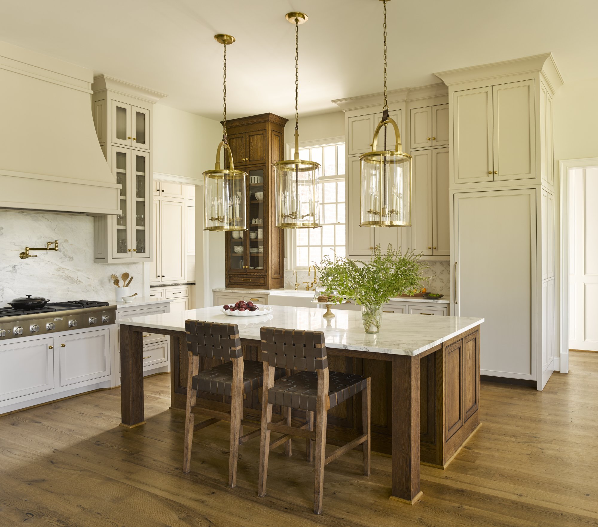 Modern kitchen with white cabinets, a kitchen island with a marble countertop, hanging pendant lights, a vase with green foliage, and wooden bar stools.