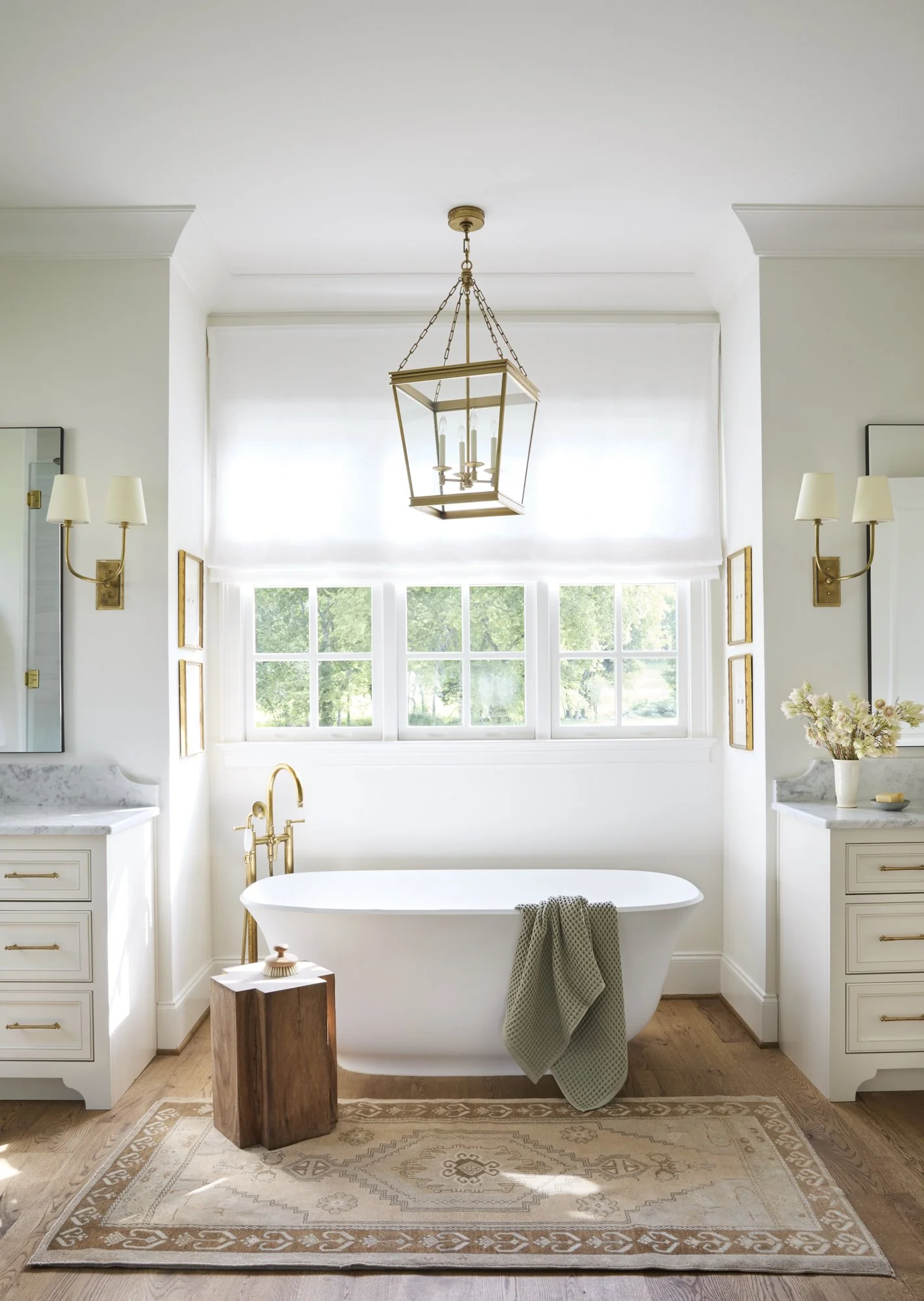 Bright bathroom with a white freestanding bathtub beneath multiple windows, gold fixtures, a beige rug, and elegant lighting fixtures on white walls.