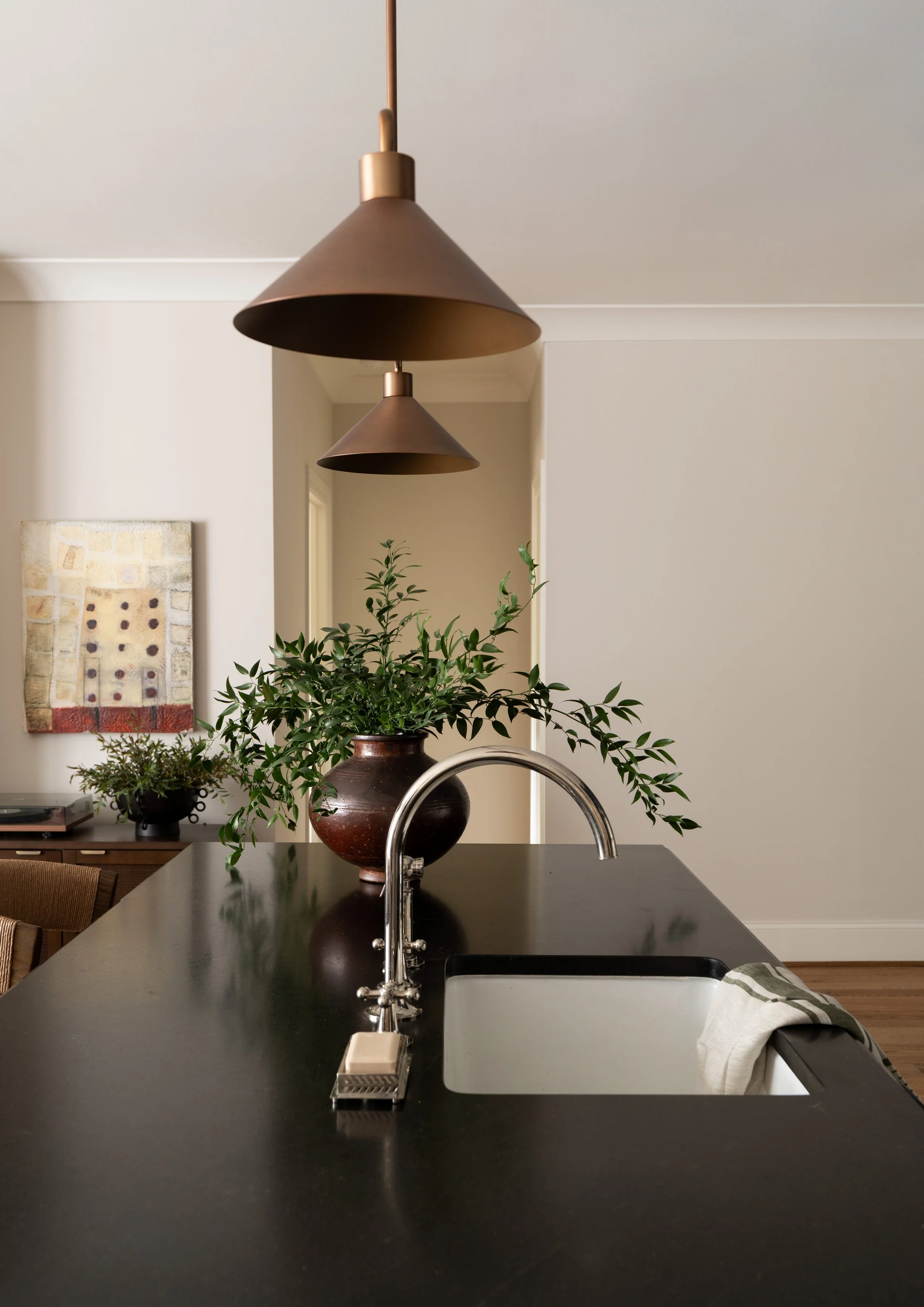 A kitchen island with a black countertop, a silver faucet, a plant in a dark brown pot, hanging brown pendant lights, and a white towel on the edge.