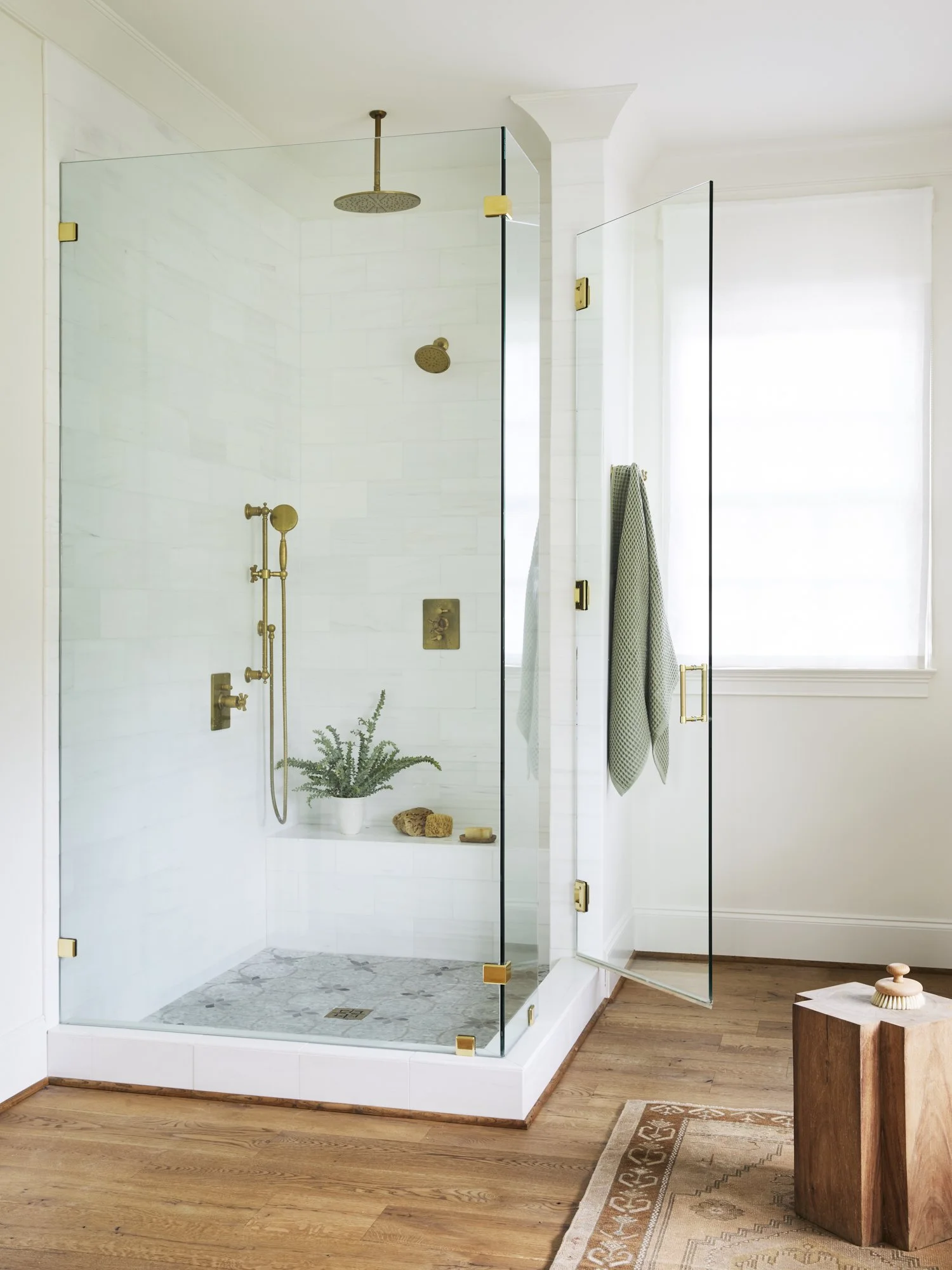 A clear glass shower enclosure with brass hardware, white tile interior, a rainfall showerhead, and a shelf with a plant and natural sponges, situated in a bright modern bathroom with wooden floor, a window with white blinds, a towel hanging on a wal