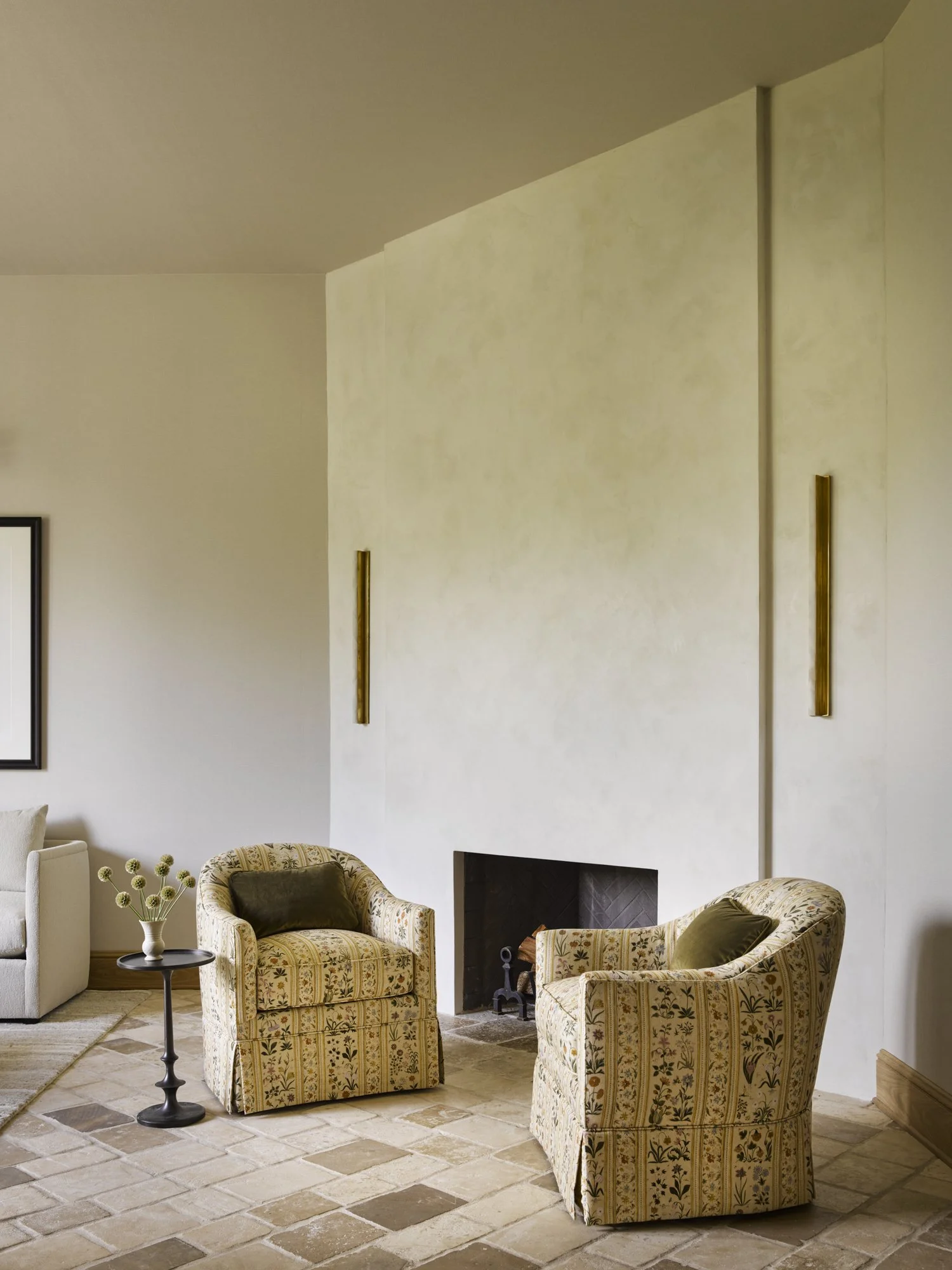 Living room with two patterned armchairs, a small black side table with a vase of flowers, a fireplace, and a beige sofa, with wood trim on the floor and gold wall-mounted decor.