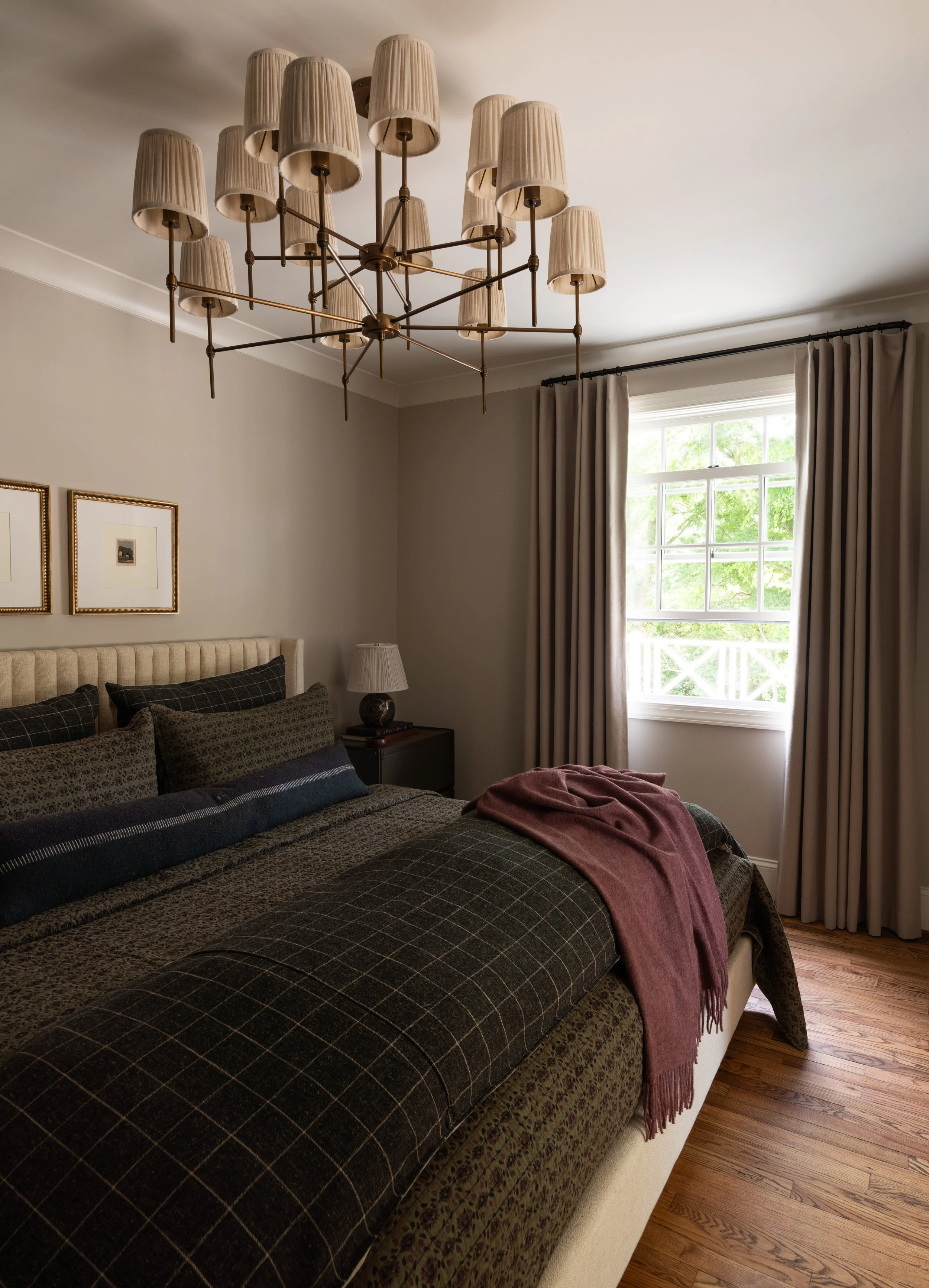 A cozy bedroom with beige walls, a tufted headboard, multiple pillows, a nightstand with a lamp, a window with curtains, a chandelier with multiple lampshades, hardwood floors, and a pink throw blanket on the bed.