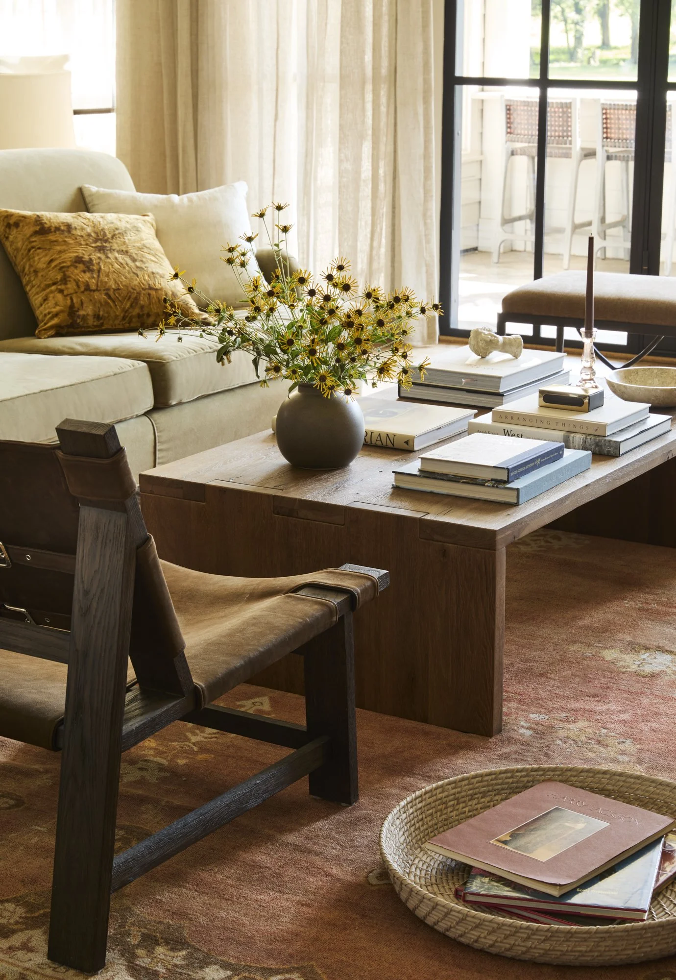 Living room with a beige sofa, a wooden coffee table with a black vase of yellow flowers, books, and a candle. A wooden chair with a leather seat is nearby, and a round woven tray on the floor contains magazines. In the background, a sliding glass do