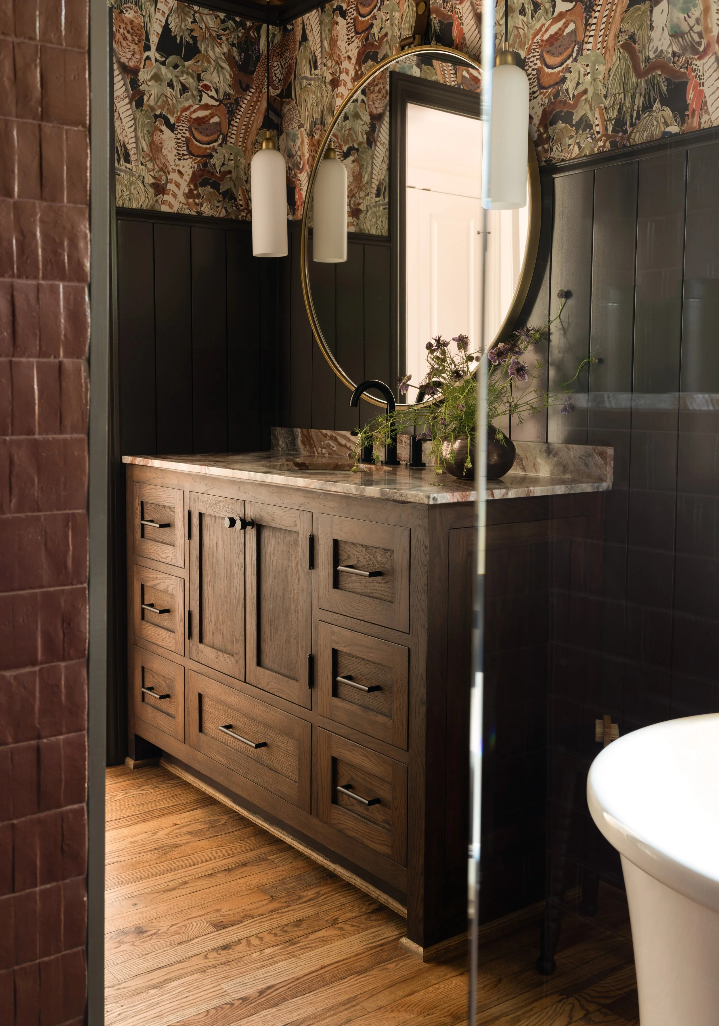 Bathroom vanity with wooden cabinet, marble countertop, circular mirror, and floral arrangement, with background wallpaper and pendant lights.