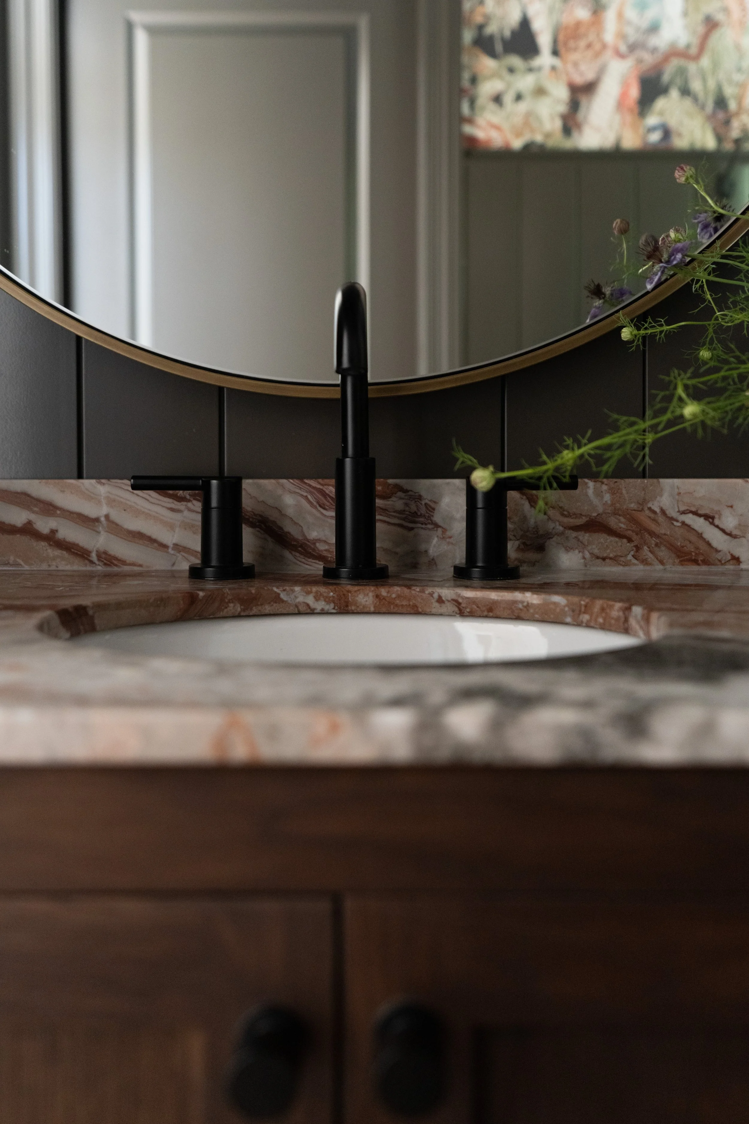 Close-up of a bathroom sink with a black faucet and a marble countertop, reflected in a circular mirror above the sink. A small plant with purple flowers is on the right side of the counter.