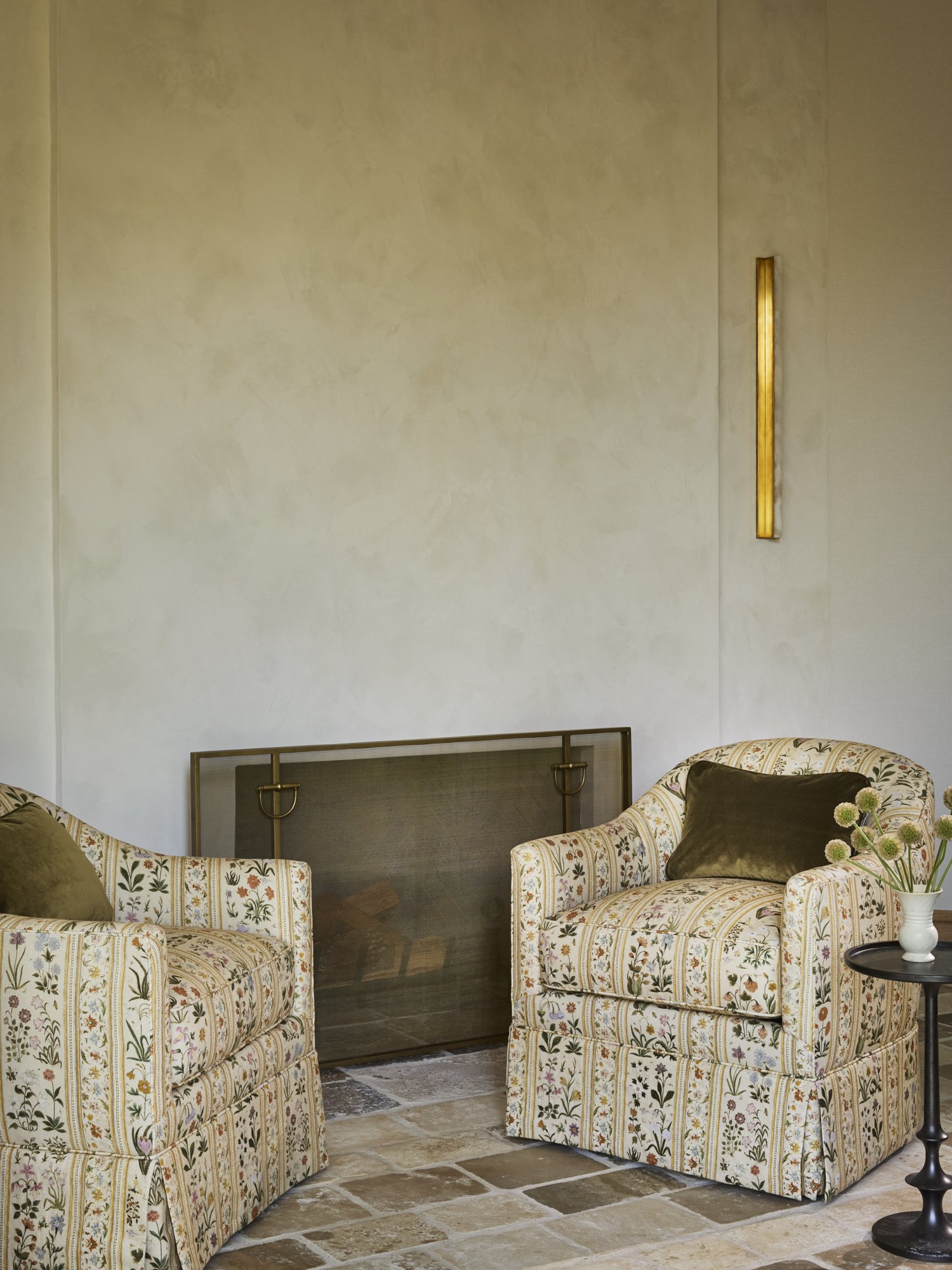 Two upholstered armchairs with floral pattern and dark green pillows, a small round black side table with a white vase and flowers, a gold wall sconce, and a fireplace with a metal screen, on a stone floor against a plain beige wall.
