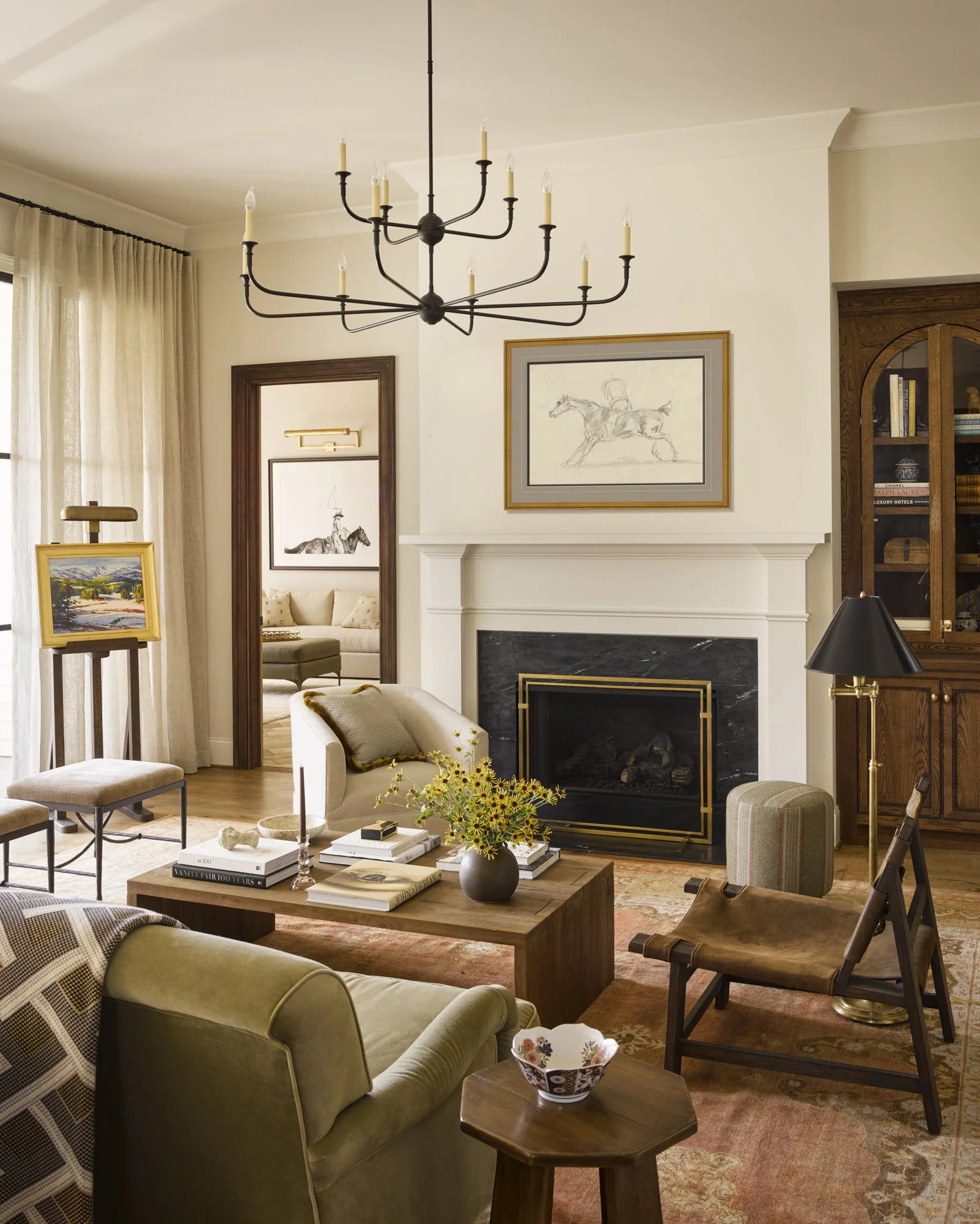 A living room with a fireplace, chandelier, armchairs, a coffee table, artwork, and a bookshelf.