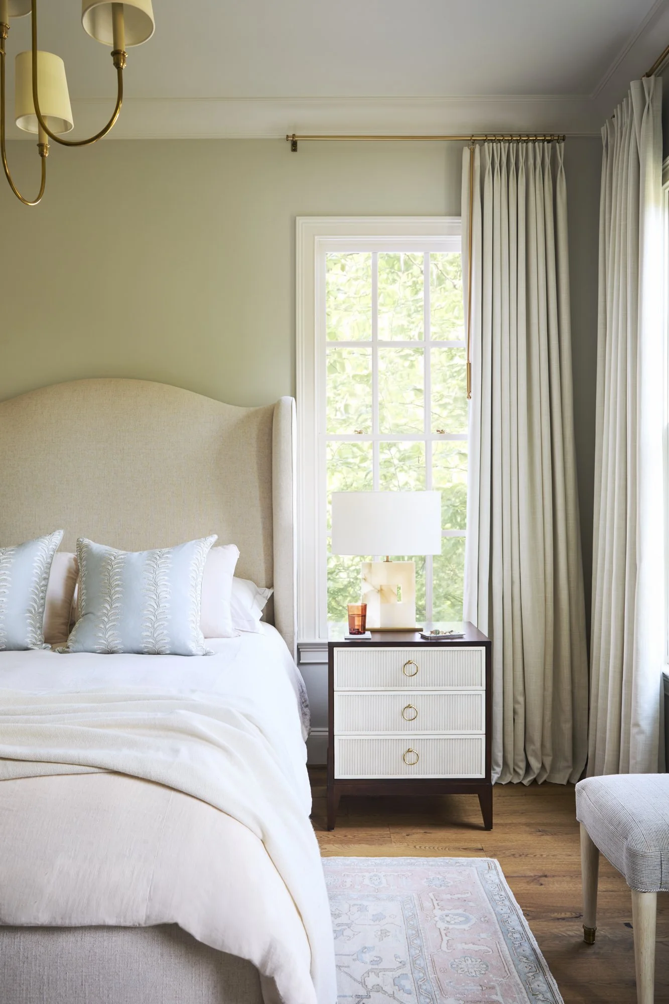 A bedroom with a large beige upholstered headboard, a window with white trim and greenery outside, cream curtains, a dark wood nightstand with white drawers and gold handles, a white lamp, a small amber glass, and a beige upholstered chair with woode