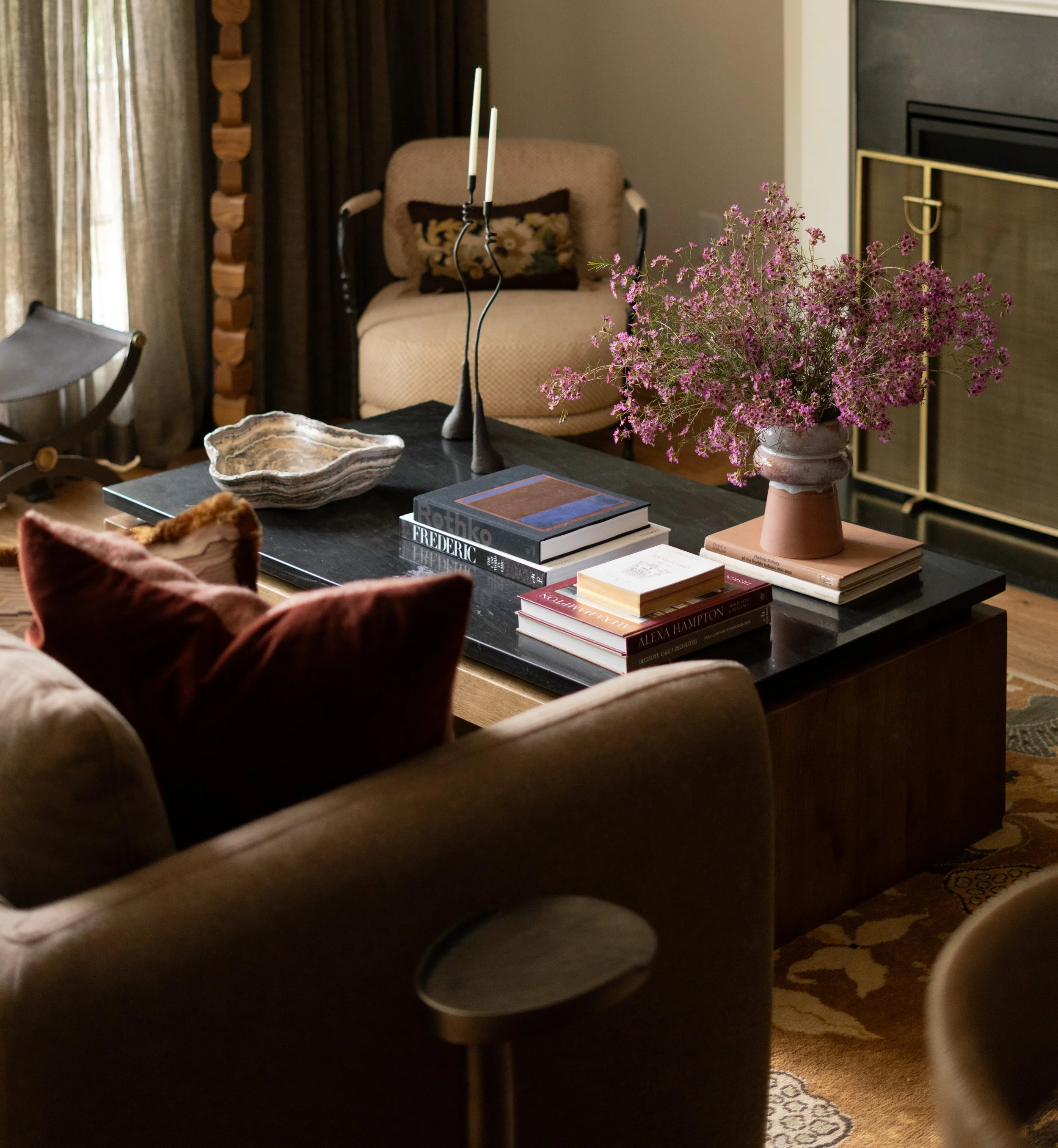 Living room with a dark wooden coffee table, a large pinkish vase with purple flowers, and various books and candles. In the background, there is a beige armchair with dark floral pillow, a fireplace with a gold-trimmed screen, and tall curtains.