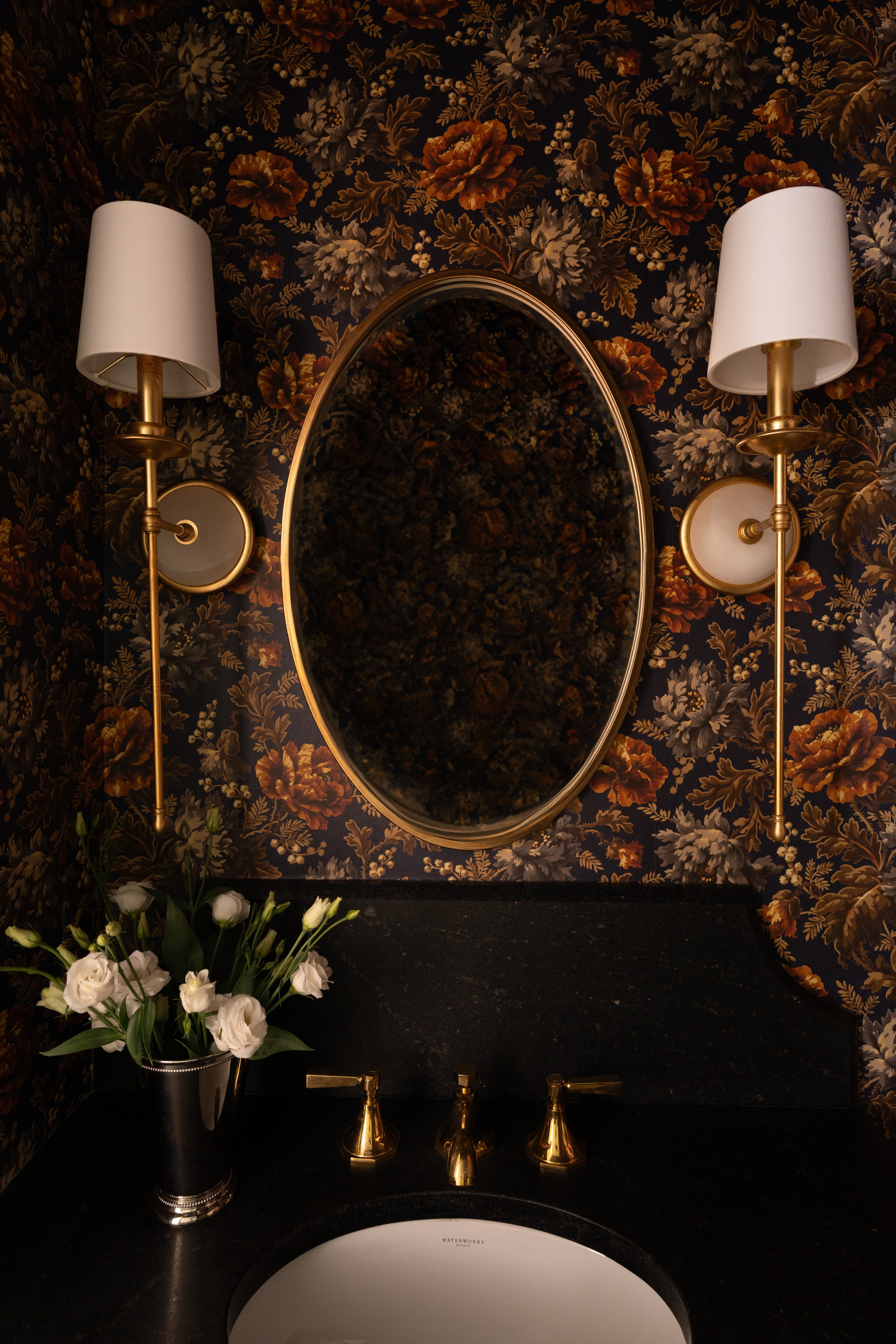 Wall-mounted mirror with gold trim flanked by two wall lamps with white shades, floral wallpaper background, black countertop with gold faucet, and a vase of white flowers.