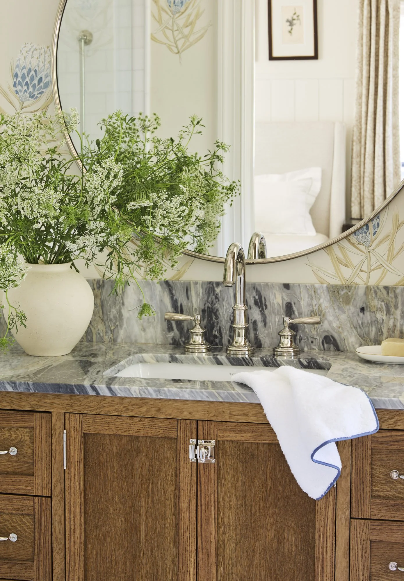 A bathroom vanity with a marble countertop, a vase with green and white flowers, a bar of soap on a dish, and a white towel hanging over the edge. A large mirror reflects part of a bed and framed artwork on the wall.