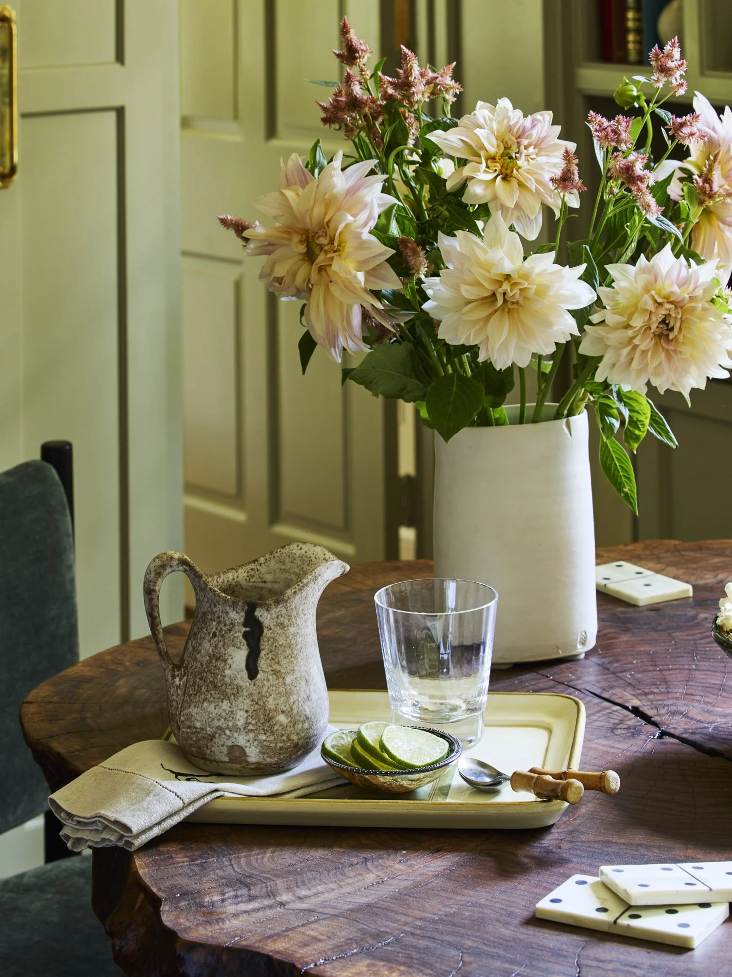 A wooden dining table with a large white vase filled with pink and cream dahlias and some greenery. On the table, there is a beige ceramic pitcher, a clear glass, a small bowl of lime slices, a spoon, and a napkin. There are also some dominoes on the