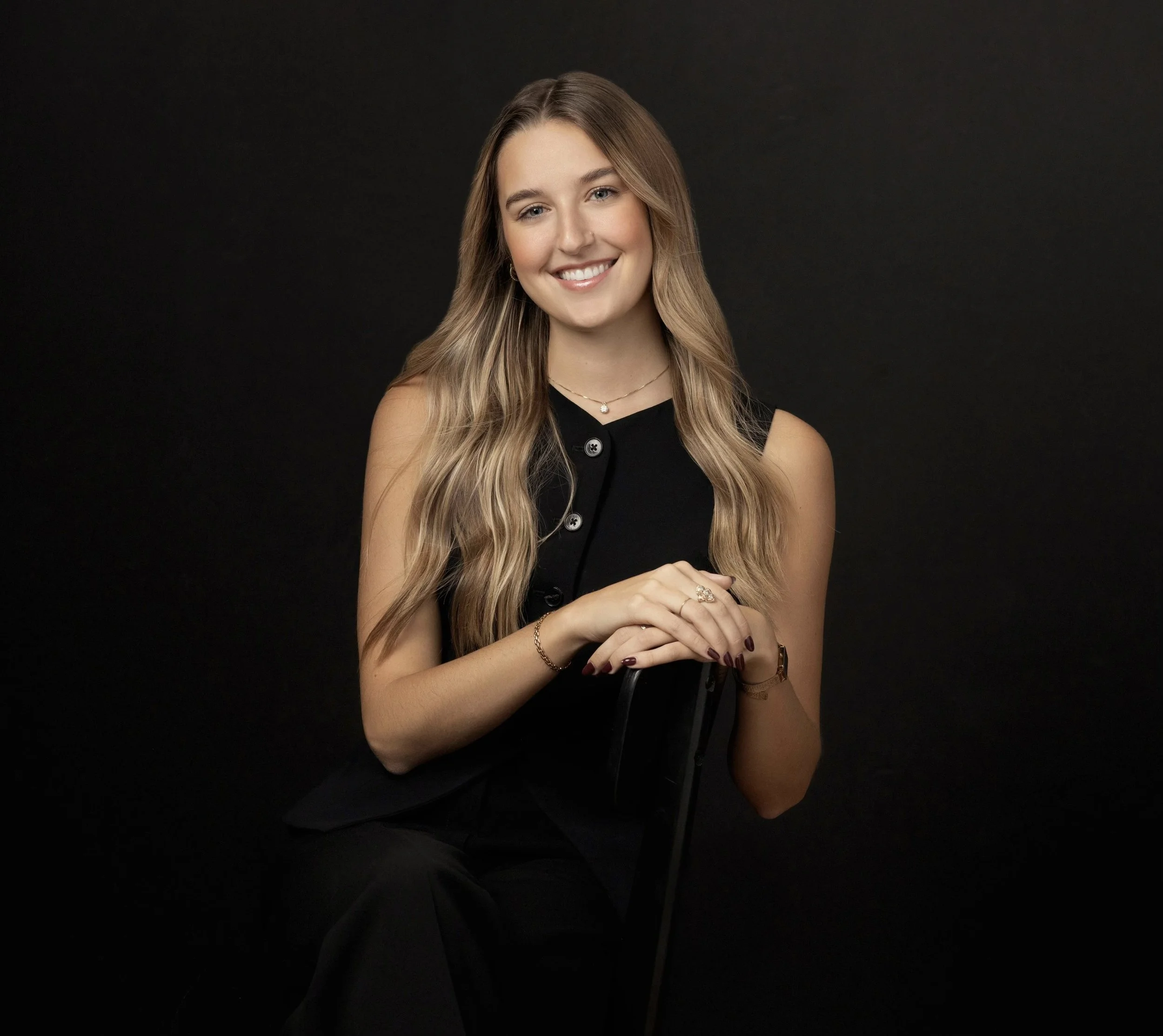 A young woman with long wavy blonde hair smiling and sitting against a black background, wearing a sleeveless black top, jewelry, and dark pants.