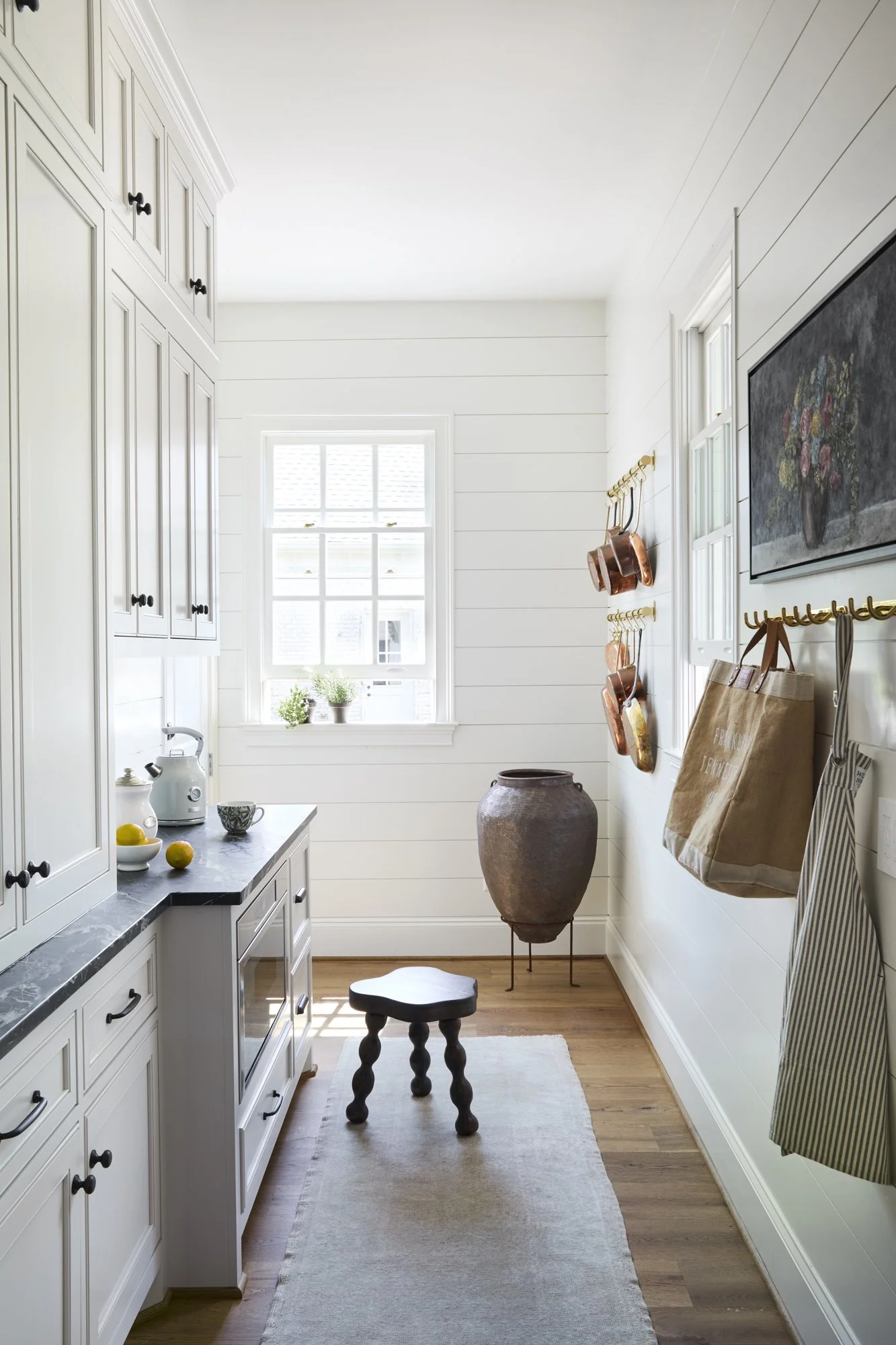 Bright kitchen with white cabinets, black countertops, a small stool, and decorative hanging copper pots. There are potted plants on the window sill and a large vase on a stand near the wall.