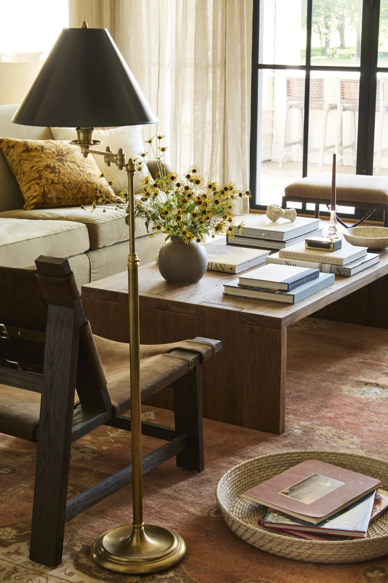Living room with a wooden coffee table holding books, a vase with yellow flowers, and a candle. A dark-colored sofa with a textured pillow, a brass floor lamp with a black shade, a sliding glass door leading to a balcony, and a rug on the floor.
