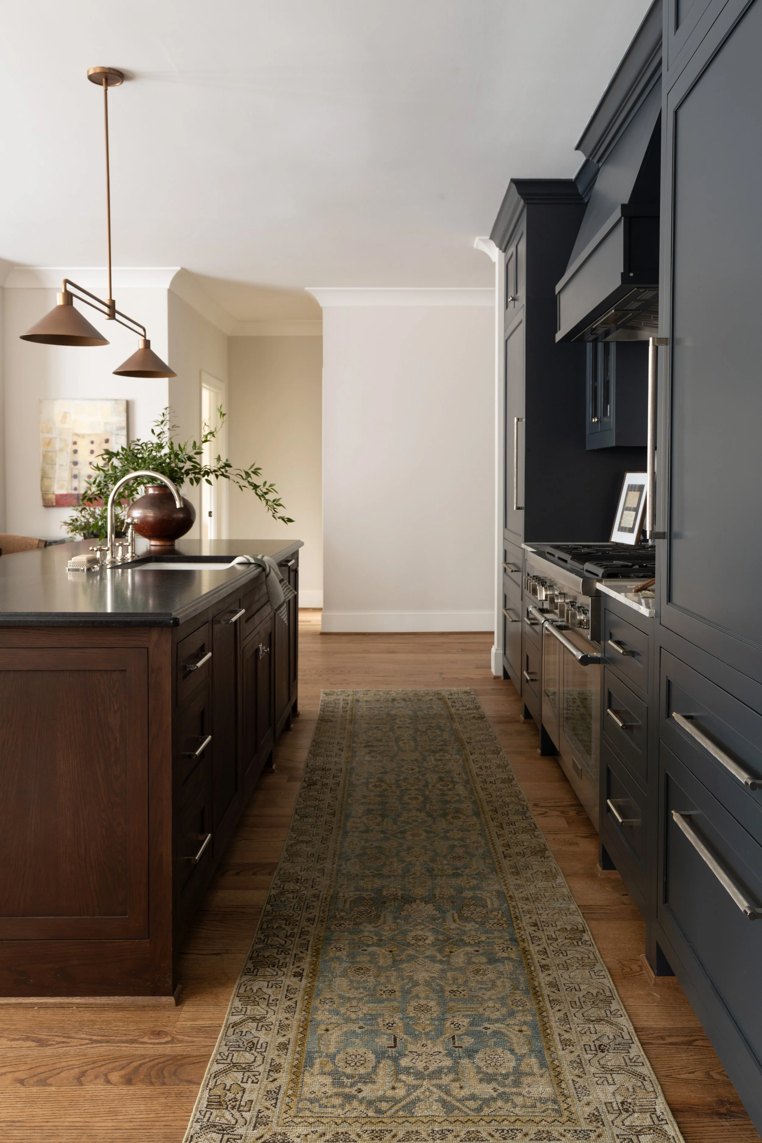A modern kitchen with a dark wood island, a black countertop, a floral arrangement in a vase, and a patterned rug on wooden floors. There are dark cabinets and stainless steel appliances along one wall, with a pendant light hanging from the ceiling.