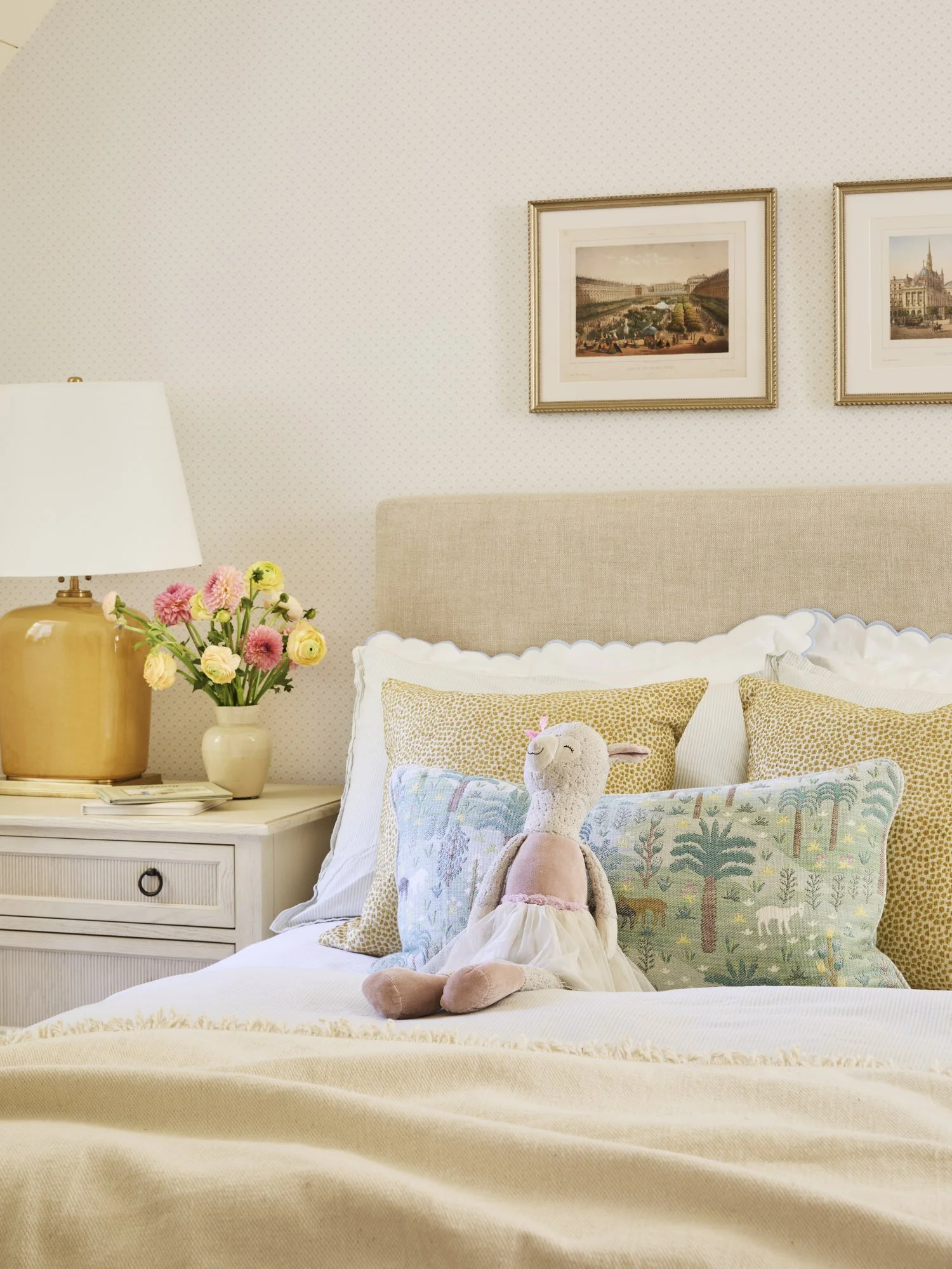 A cozy bedroom with a bed that has a plush llama stuffed animal dressed in a tutu, decorative pillows with nature and animal prints, a nightstand with a vase of pink and yellow flowers, a lamp, and framed pictures of scenic landscapes on the wall.