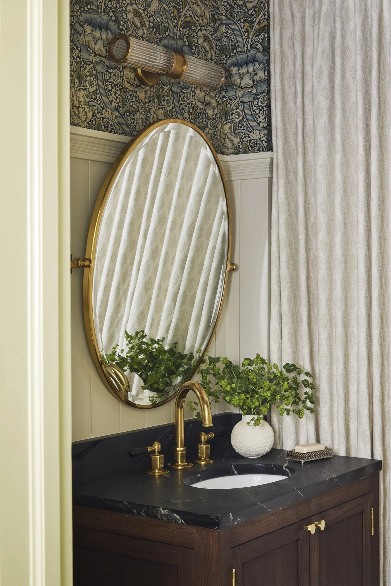 Bathroom vanity with a black marble countertop, gold fixtures, a white textured vase with greenery, a small soap dish, an oval mirror with a gold frame, beige patterned curtains, and wallpaper with blue and gold floral design.