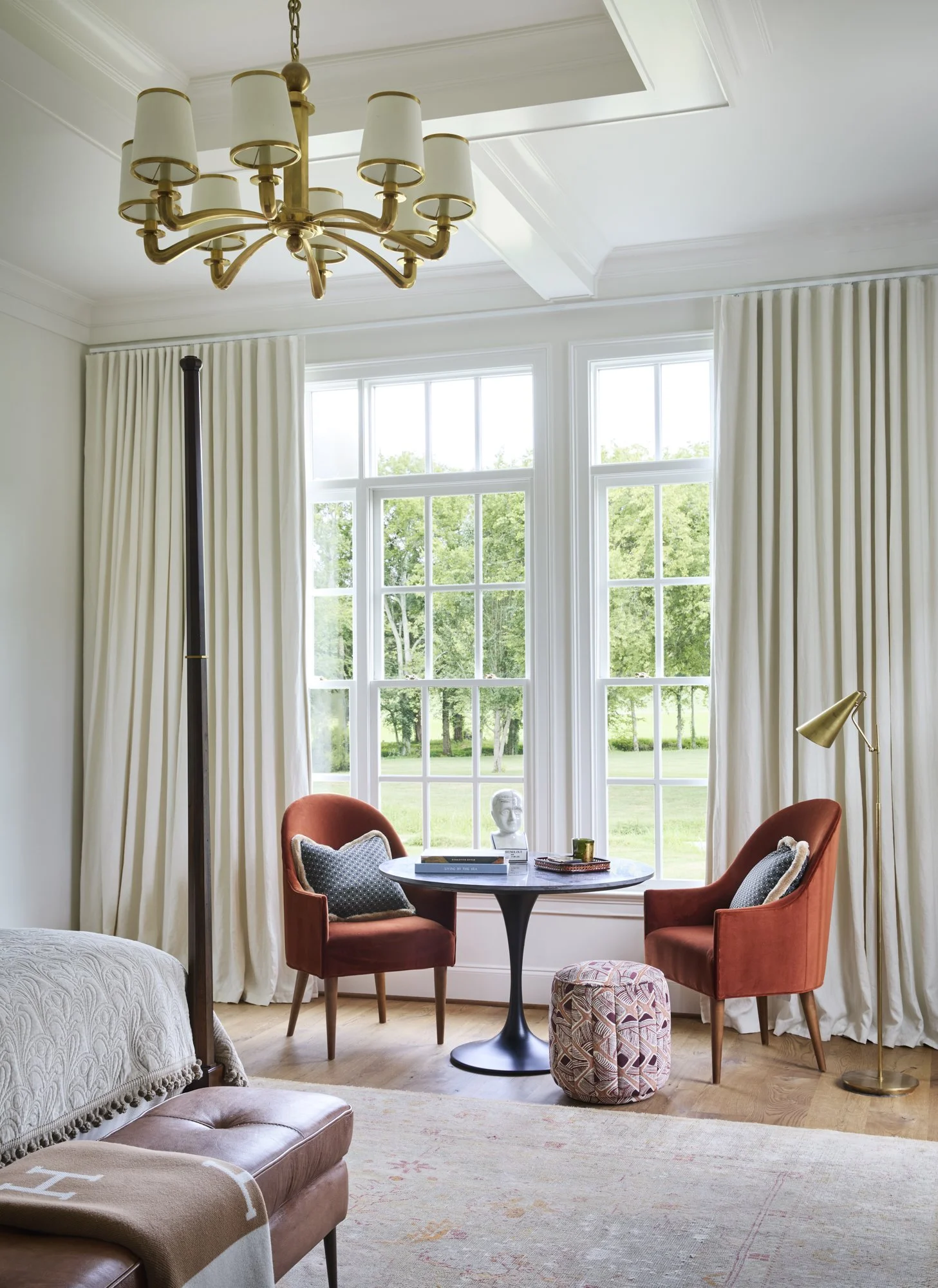 Interior view of a bright, elegant bedroom with large windows, white curtains, a gold chandelier, and cozy seating area with two orange armchairs and a round table.