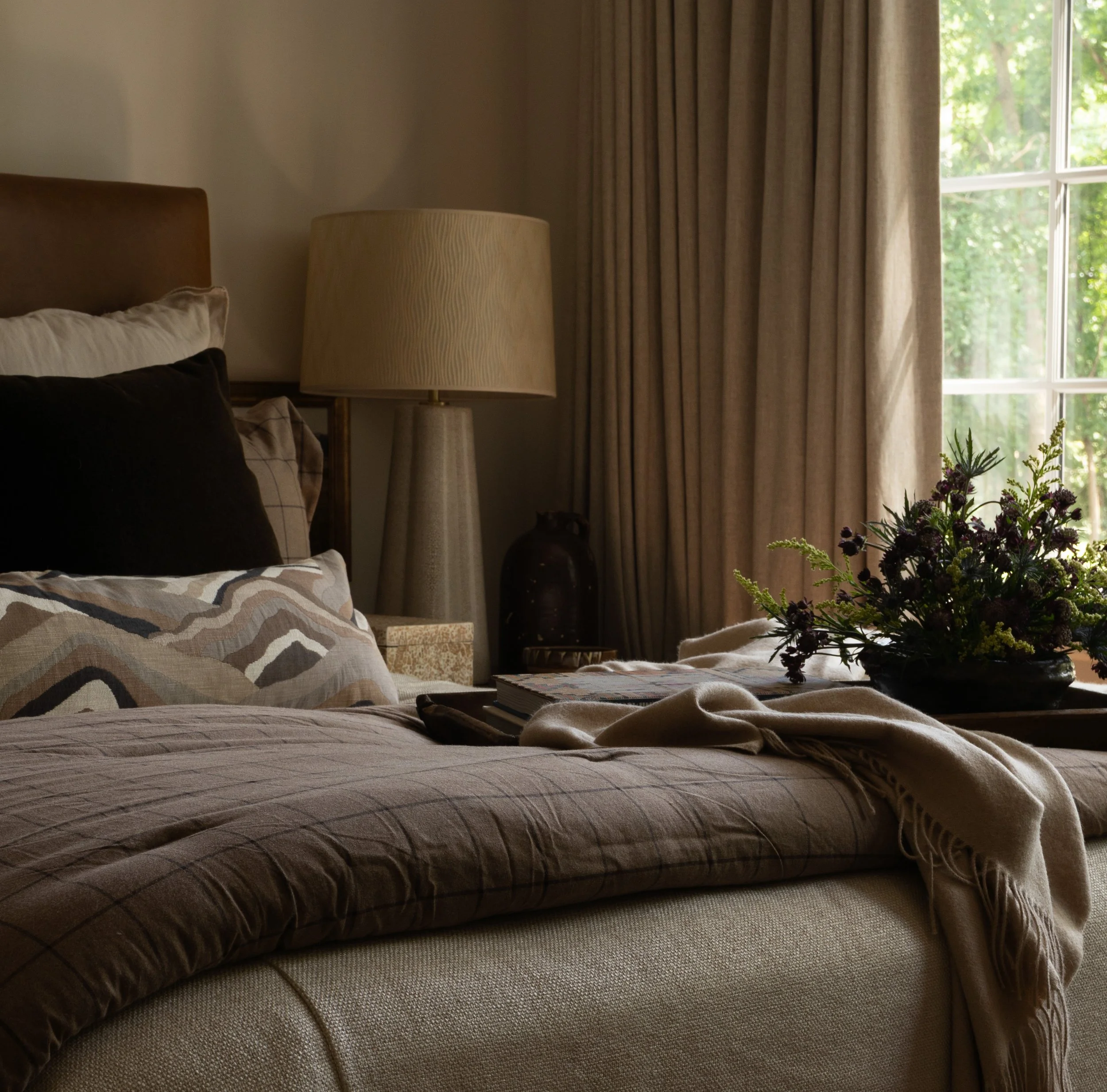 A cozy bedroom with a bed decorated with patterned and black pillows, a beige blanket, a bedspread, and an open book on the bed. There is a large window with beige curtains, a tall beige lampshade, a dark vase, and a vase of purple flowers on a side