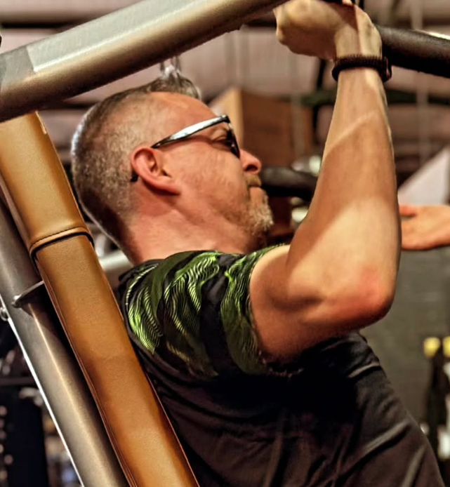Man with sunglasses performs a pull-up on gym equipment inside a fitness center.