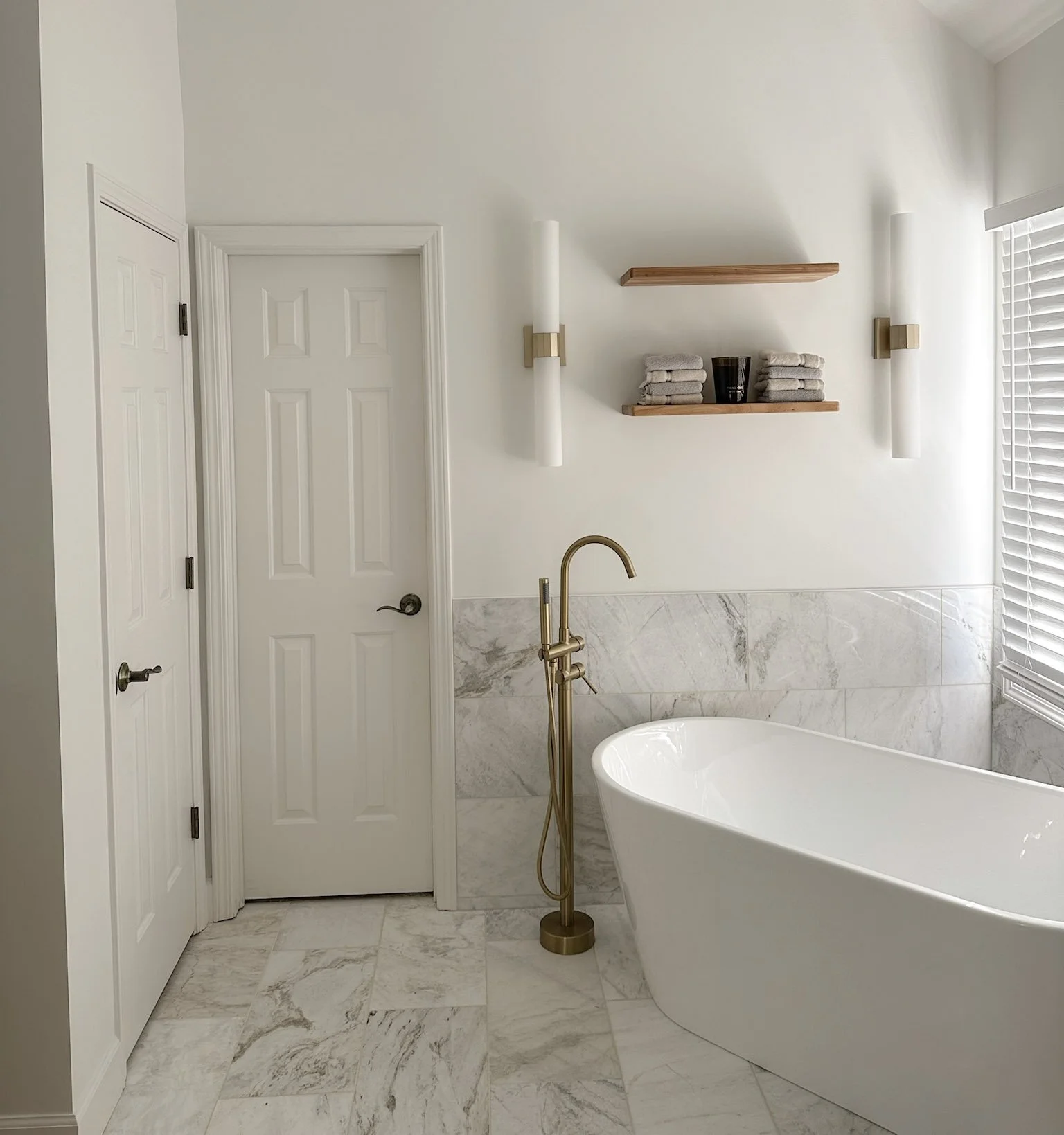 Modern bathroom with freestanding tub, marble-look tile flooring, brass floor-mounted faucet, and open wall shelving.
