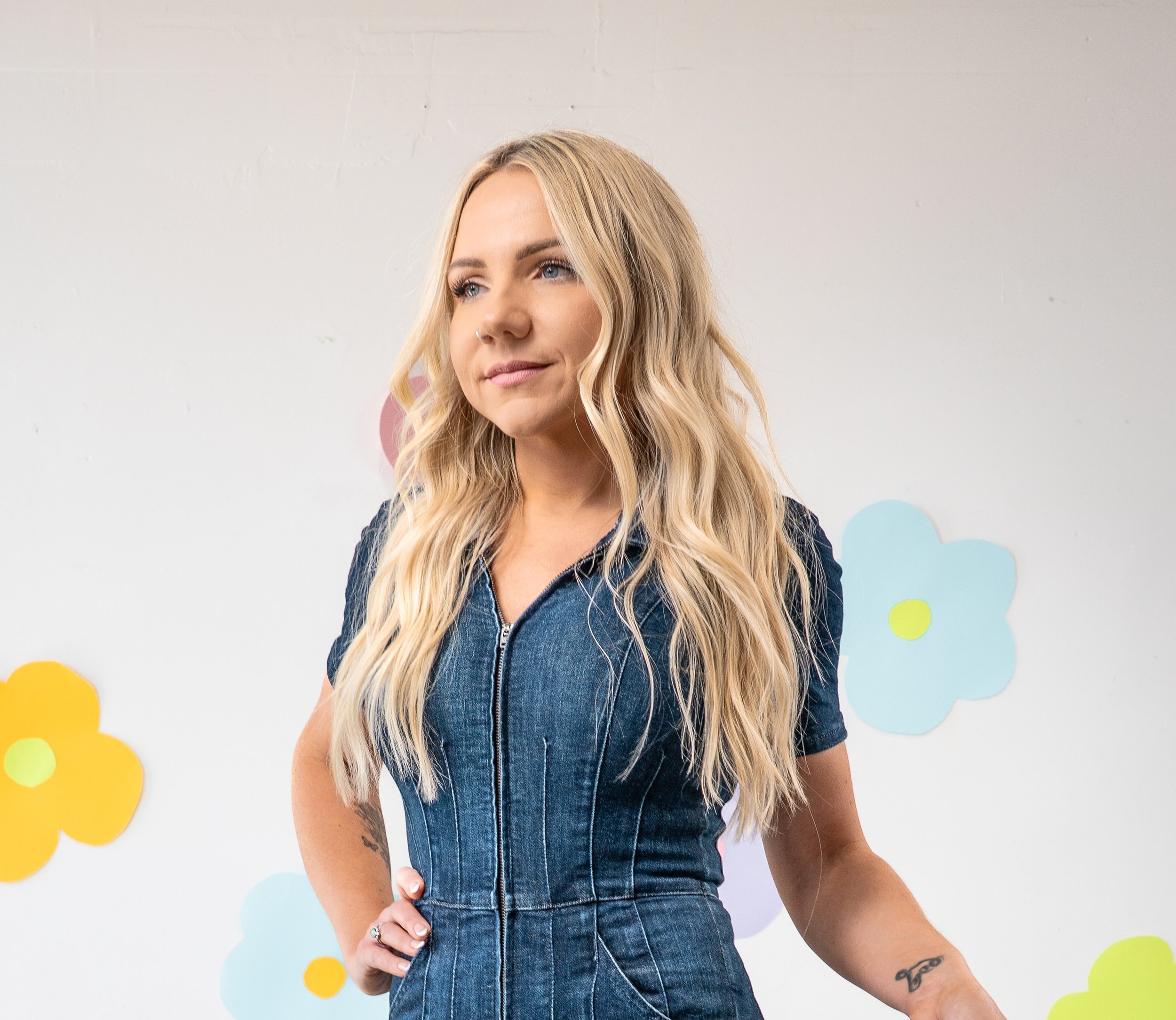 Danielle from Honey Bee Color Collective with long blonde waves, wearing a denim jumpsuit in front of a daisy-print wall.