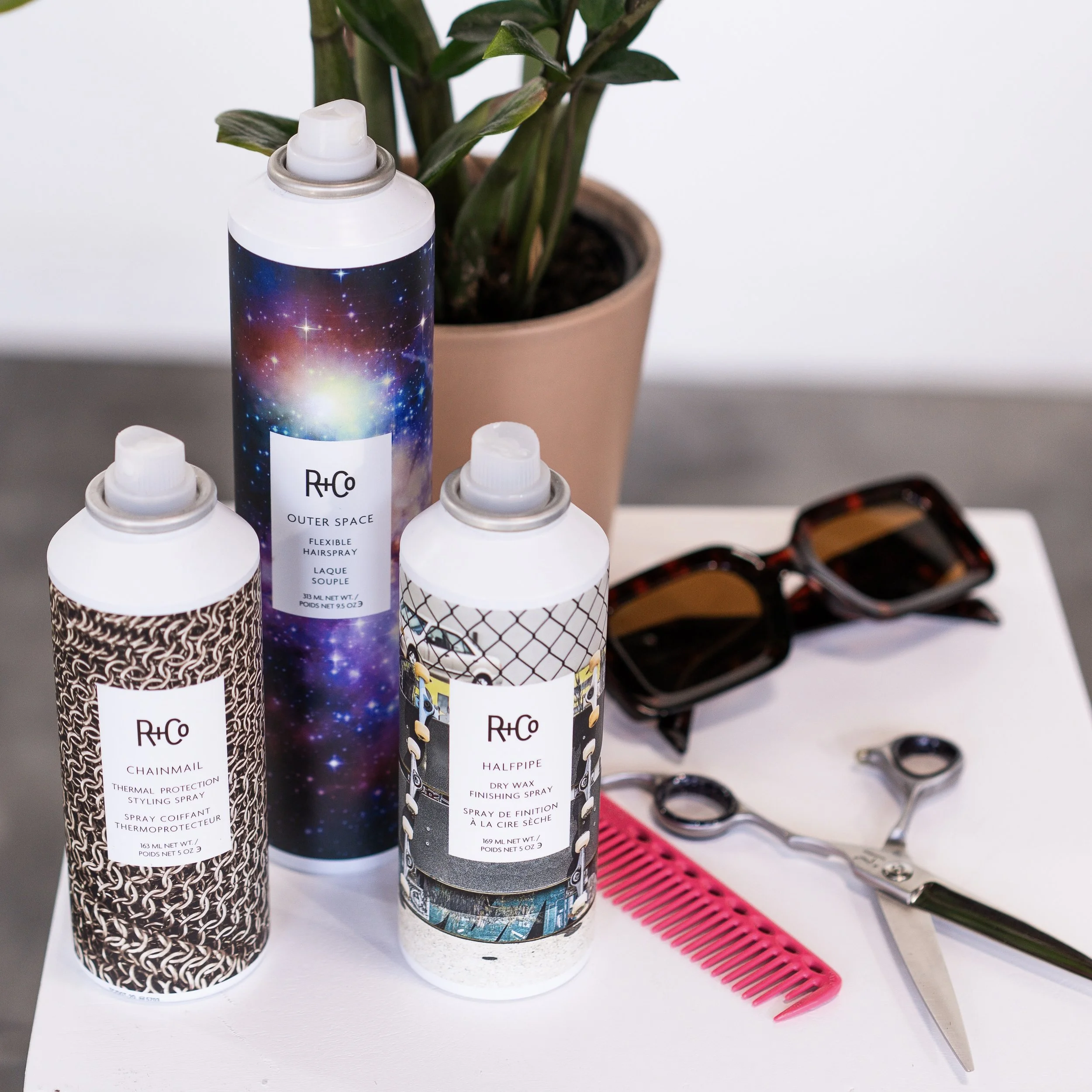 Three R+Co hair styling products, a pink comb, shears, and sunglasses arranged on a white table with a potted plant.