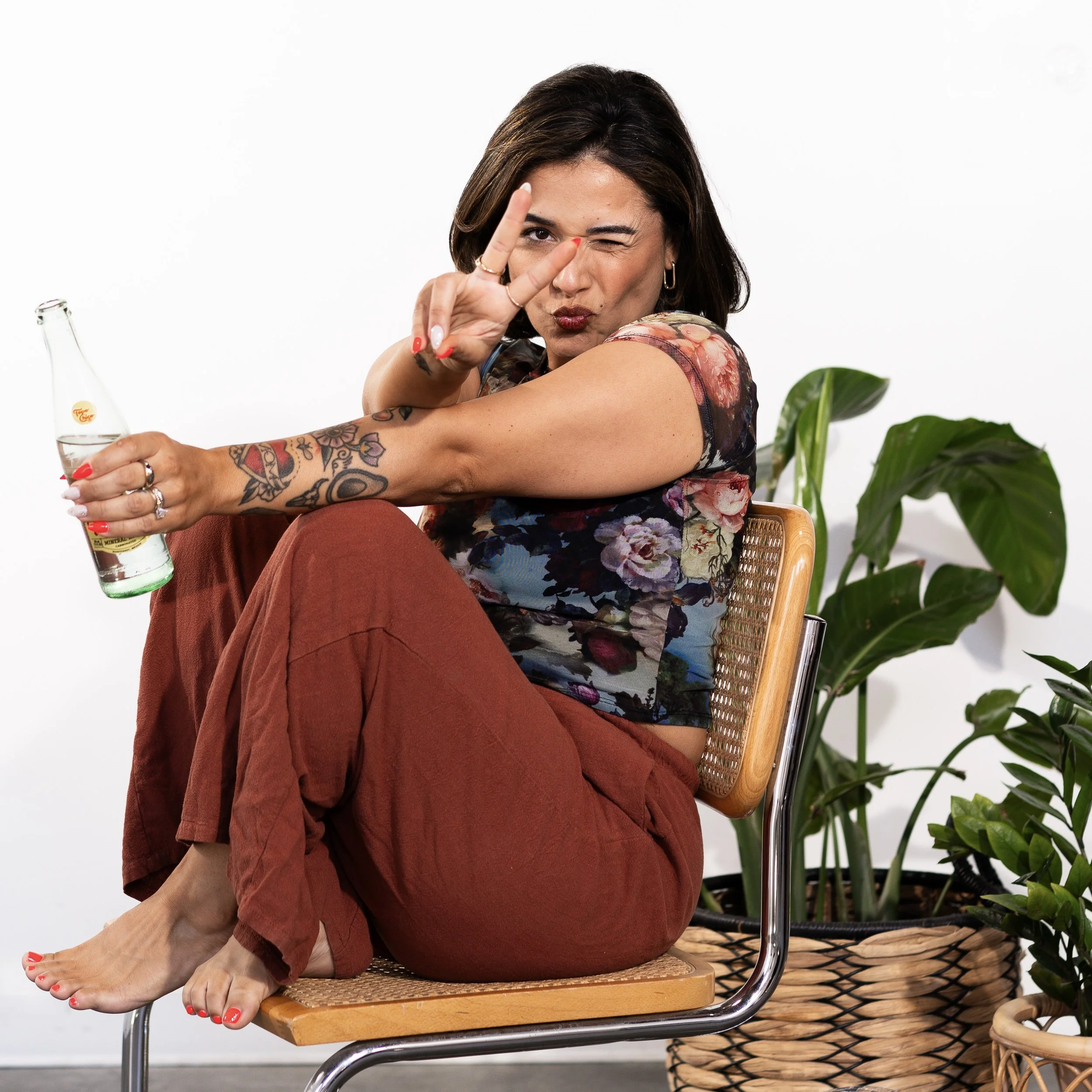 Maria from Honey Bee Color Collective sitting playfully on a chair, holding a bottle and making a peace sign, surrounded by potted plants.