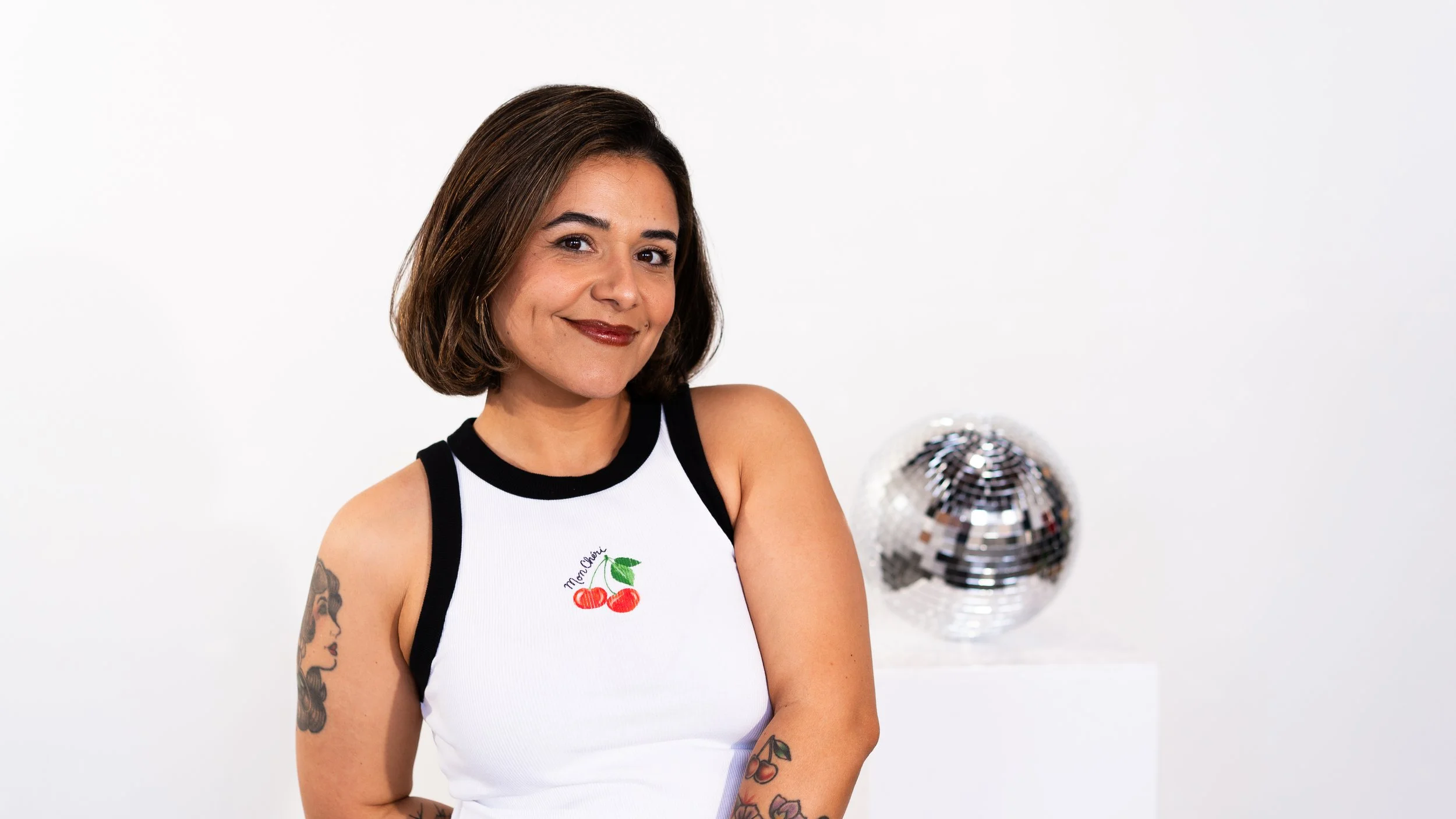 Maria from Honey Bee Color Collective, smiling with short brown hair, wearing a white and black tank top, with a disco ball in the background.