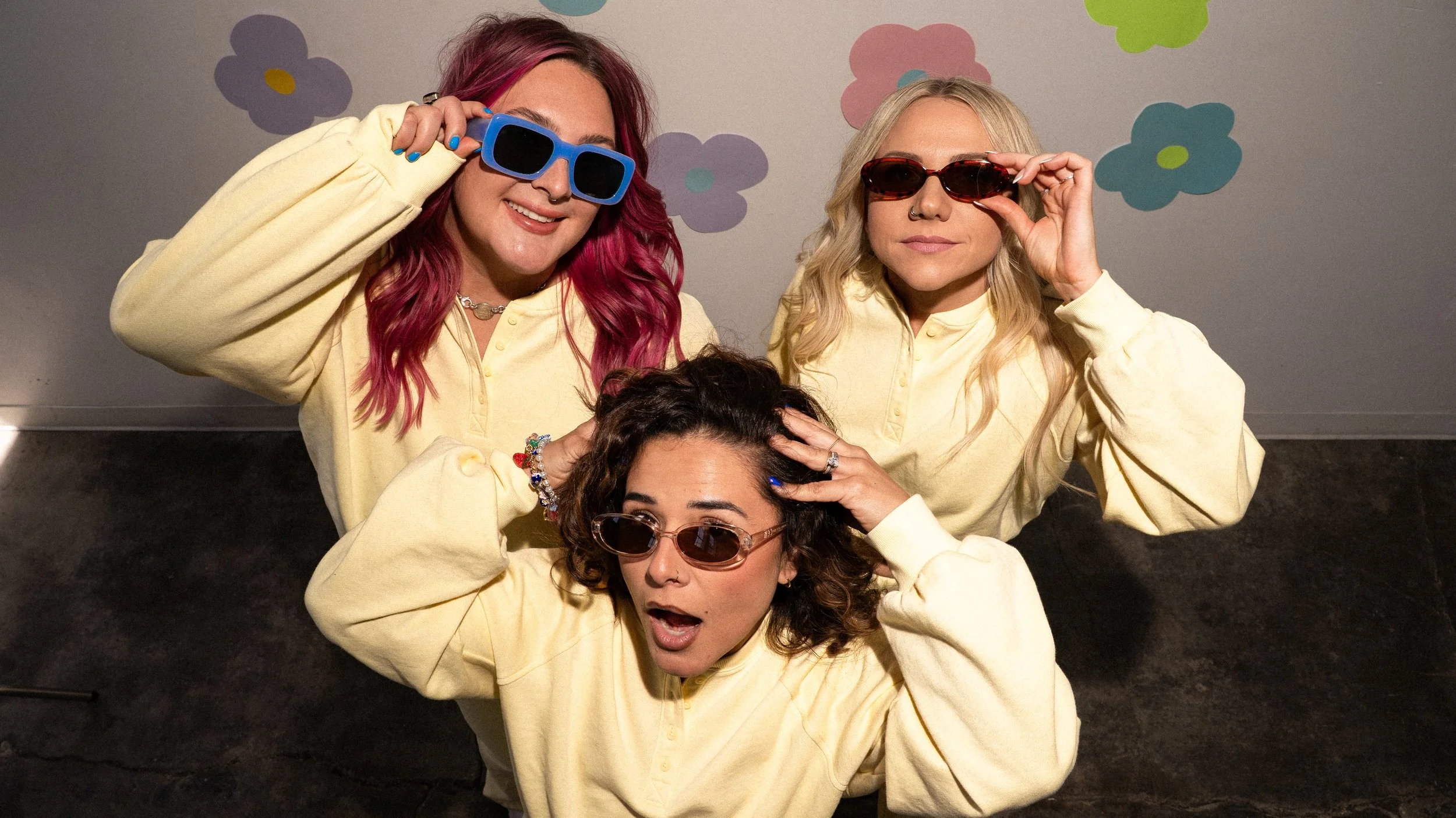 Three stylists from Honey Bee Color Collective in matching pale yellow sweatshirts and sunglasses, playfully posing in front of a floral wall.