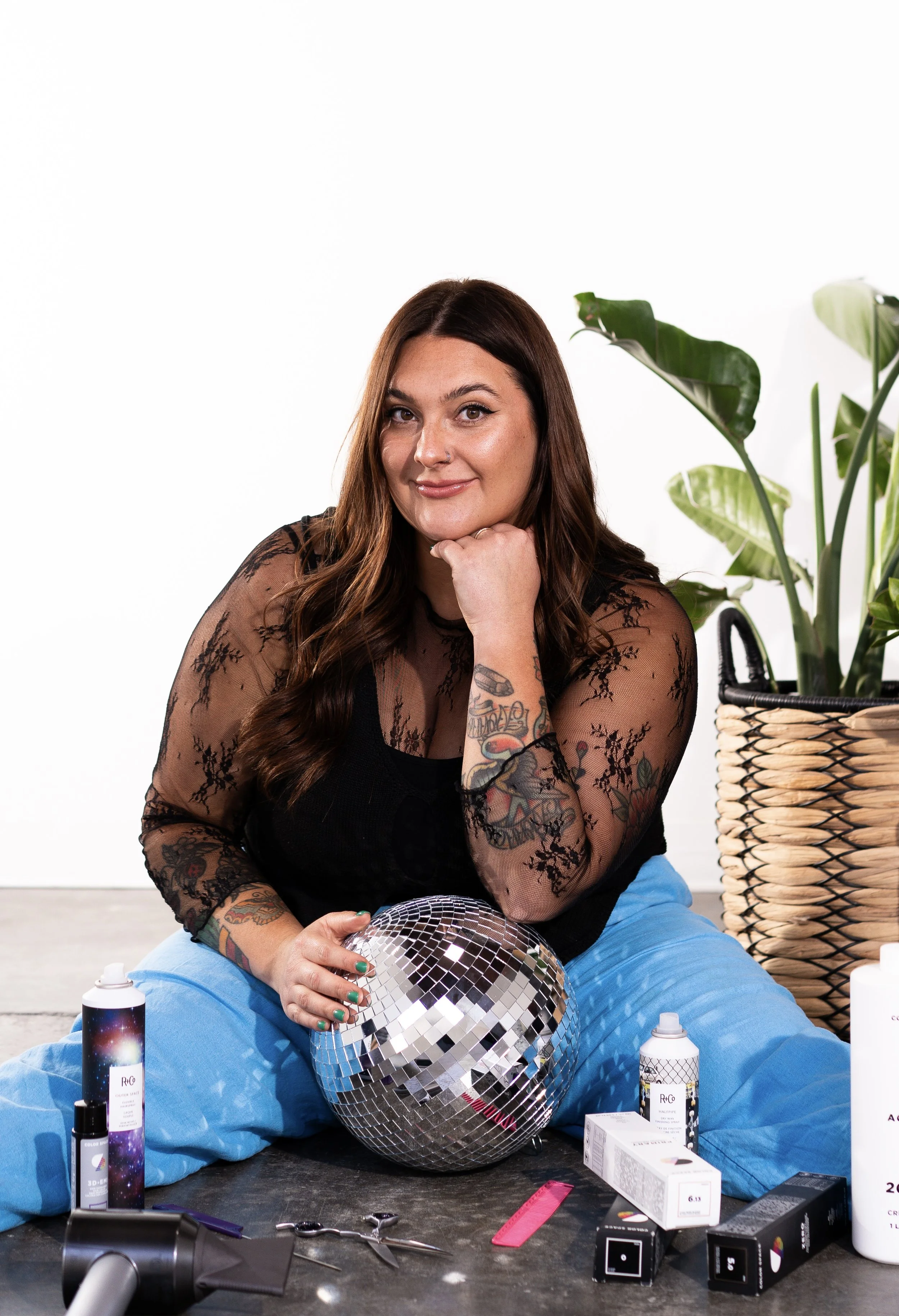 Cid from Honey Bee Color Collective with long brown hair, resting her chin on her hand and holding a disco ball, seated next to a potted plant.