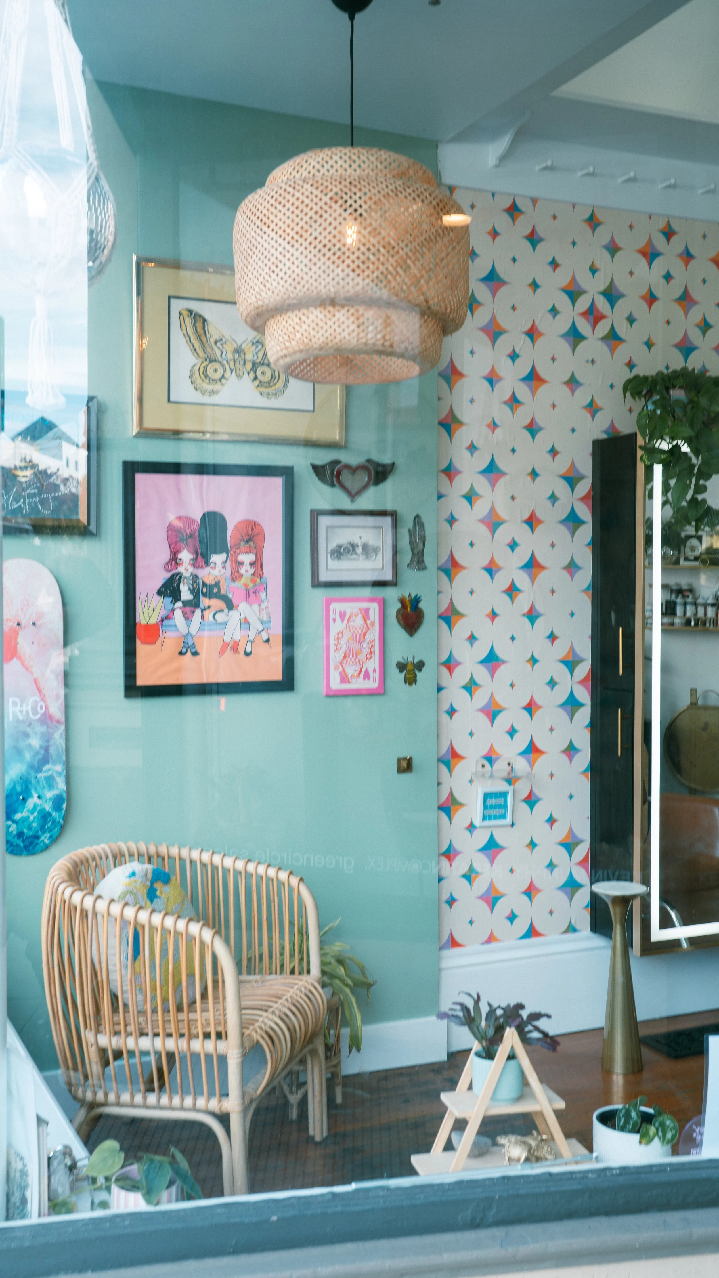 A cozy, eclectic salon interior at Honey Bee Color Collective, with a mint green wall covered in colorful art, a woven rattan lamp and chair, patterned wallpaper, and potted plants.