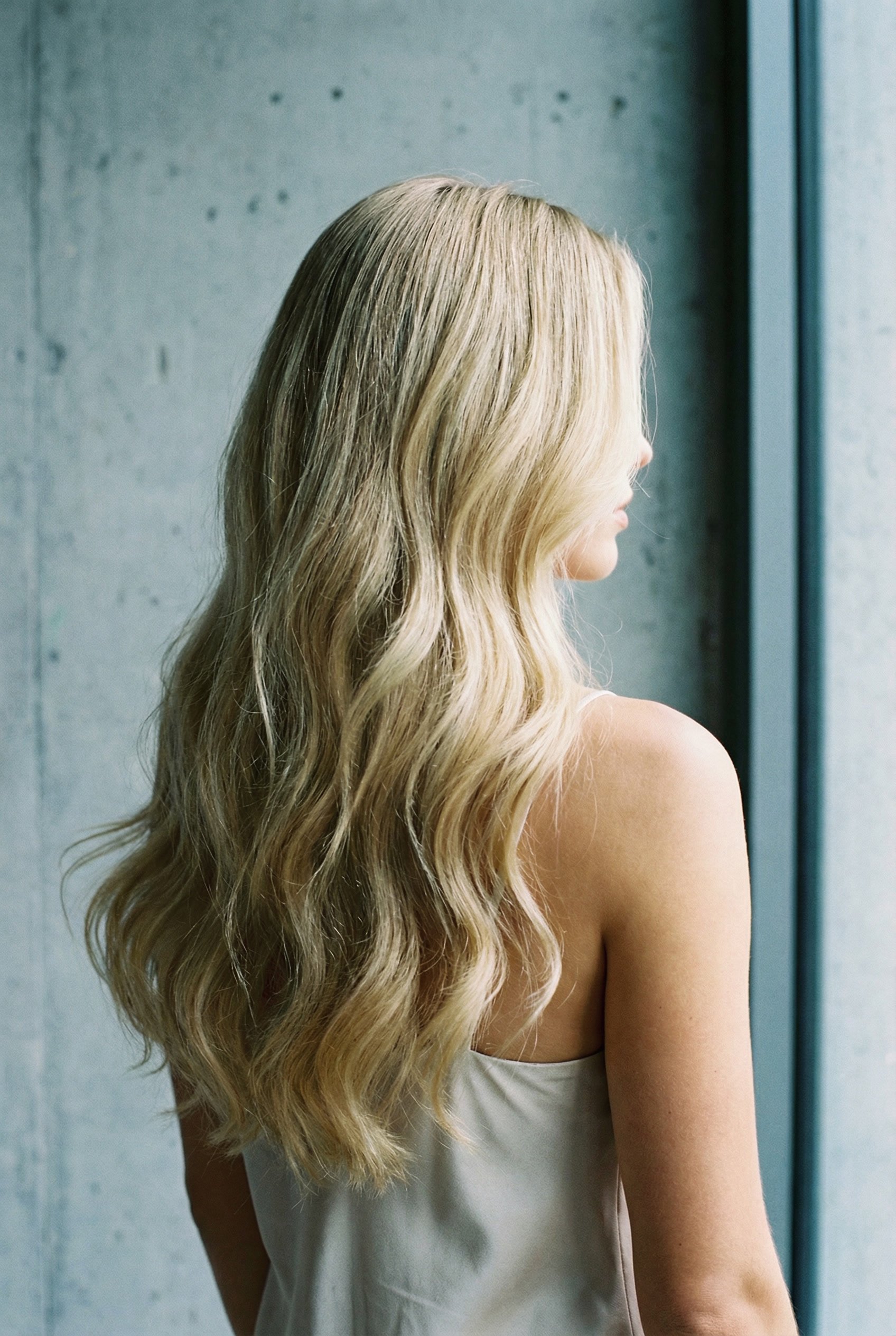 A woman with long blonde hair and blue eyes wearing a white shirt, standing indoors near a window.