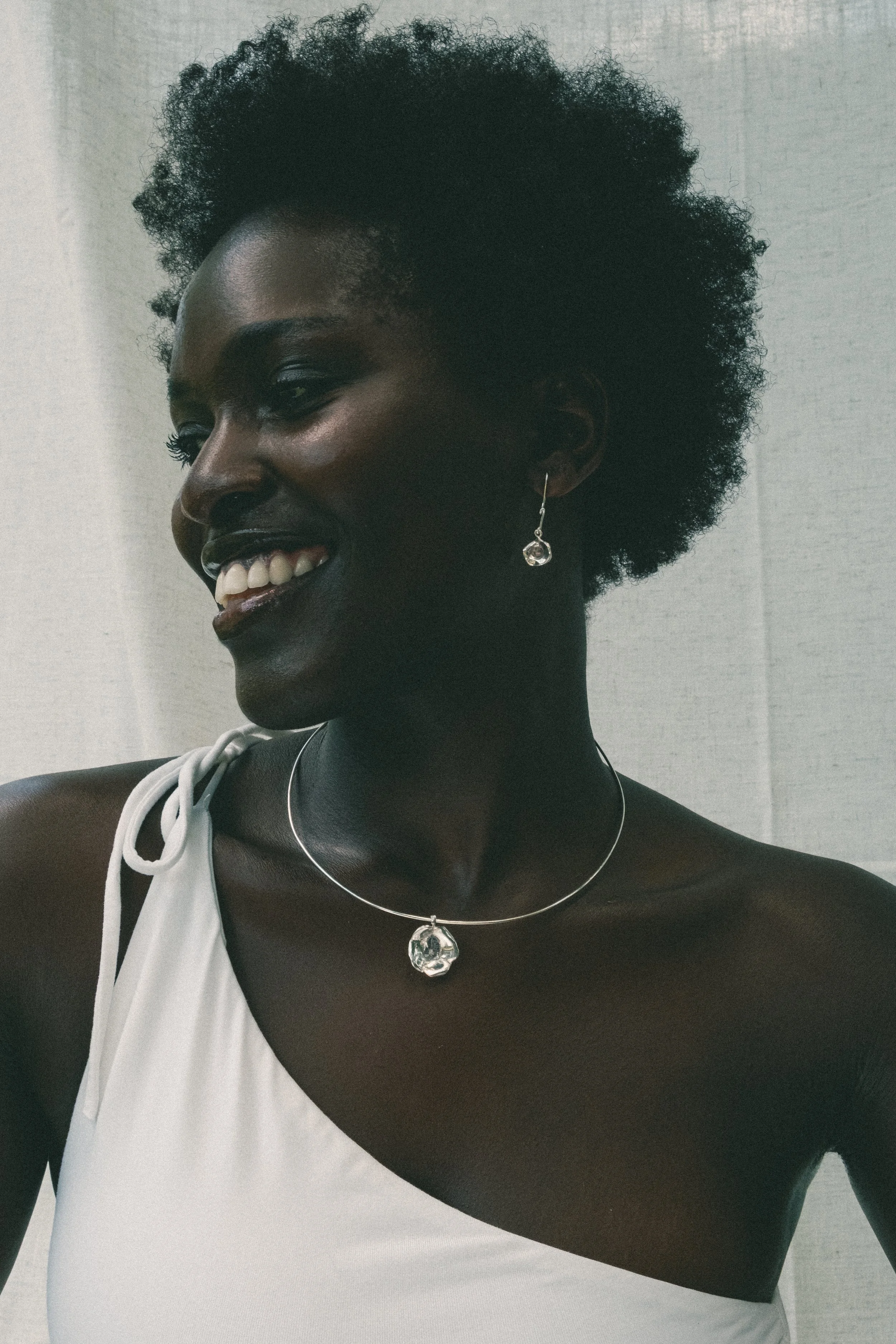 Portrait of a smiling Black woman with short, curly hair, wearing a white top, silver jewelry, and earrings, standing against a plain background.