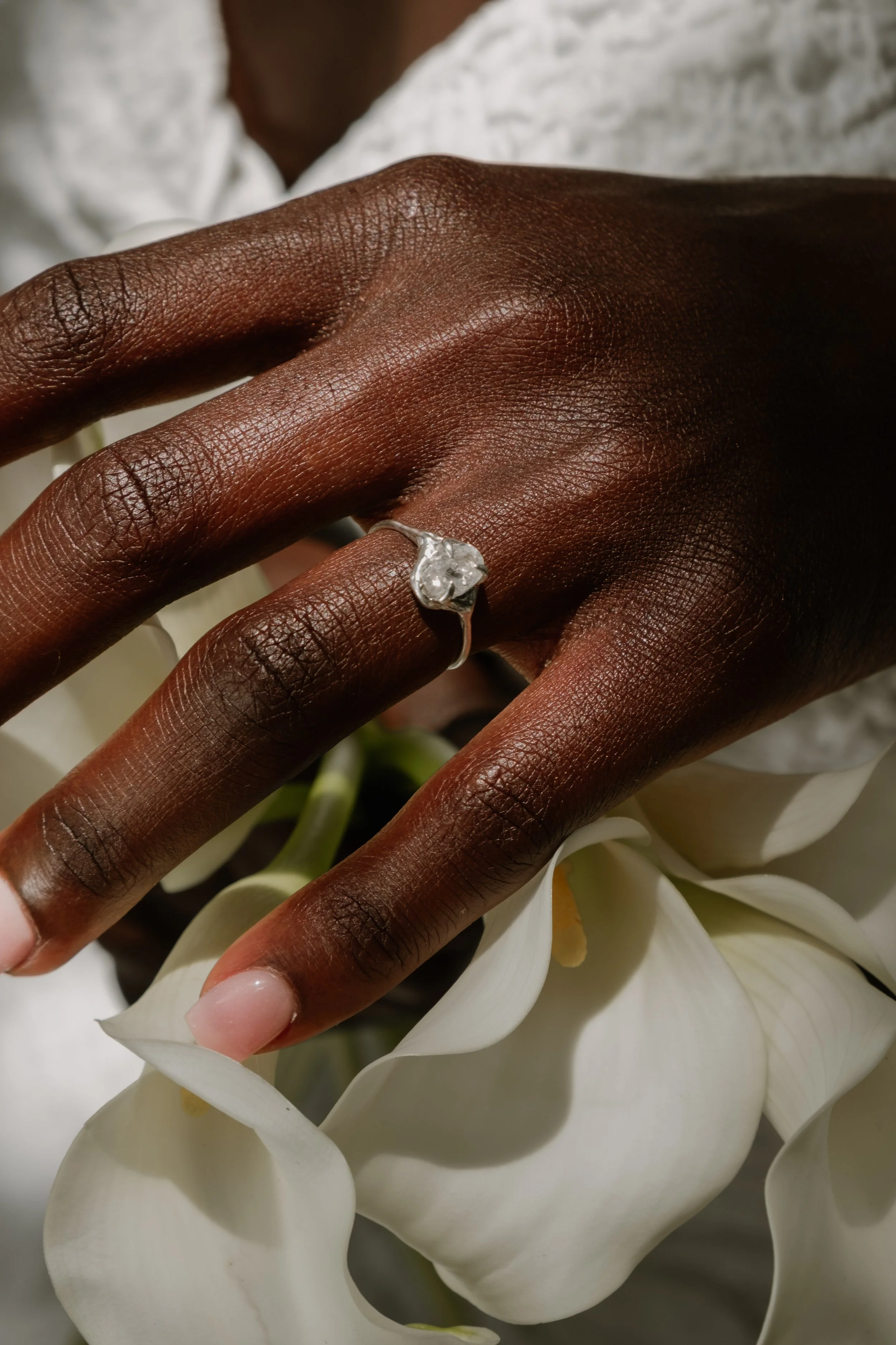 A hand with dark skin wearing a white gold ring with a large gemstone, resting on white calla lily flowers.
