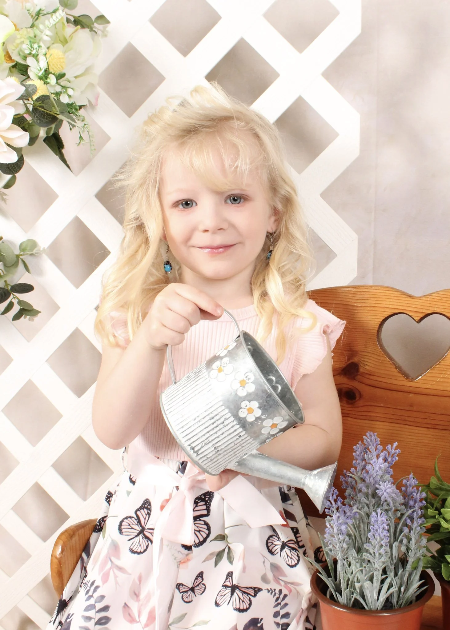 A young girl with blonde hair, wearing a pink top and a butterfly-patterned skirt, smiling and holding a small metal watering can with white and orange flowers painted on it. She is sitting on a wooden bench with a heart-shaped cutout and surrounded by potted plants, with a white lattice and flowers in the background.