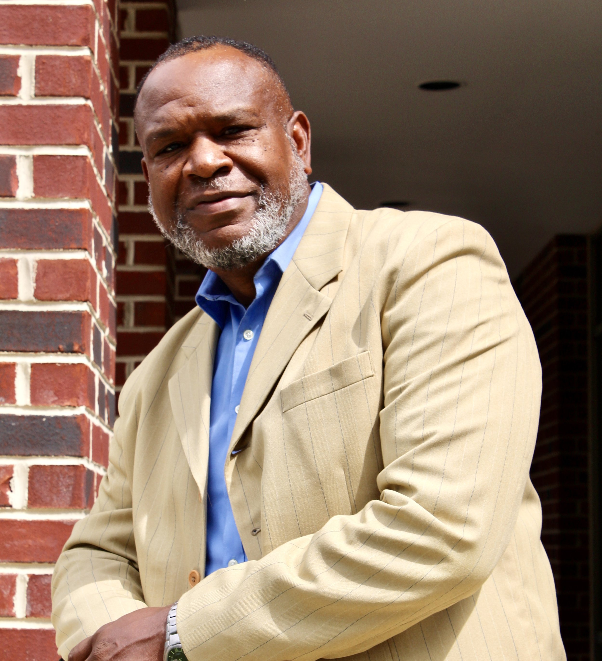 An older African American man with gray beard and short hair, wearing a light-colored blazer and blue shirt, standing outdoors next to a brick wall.